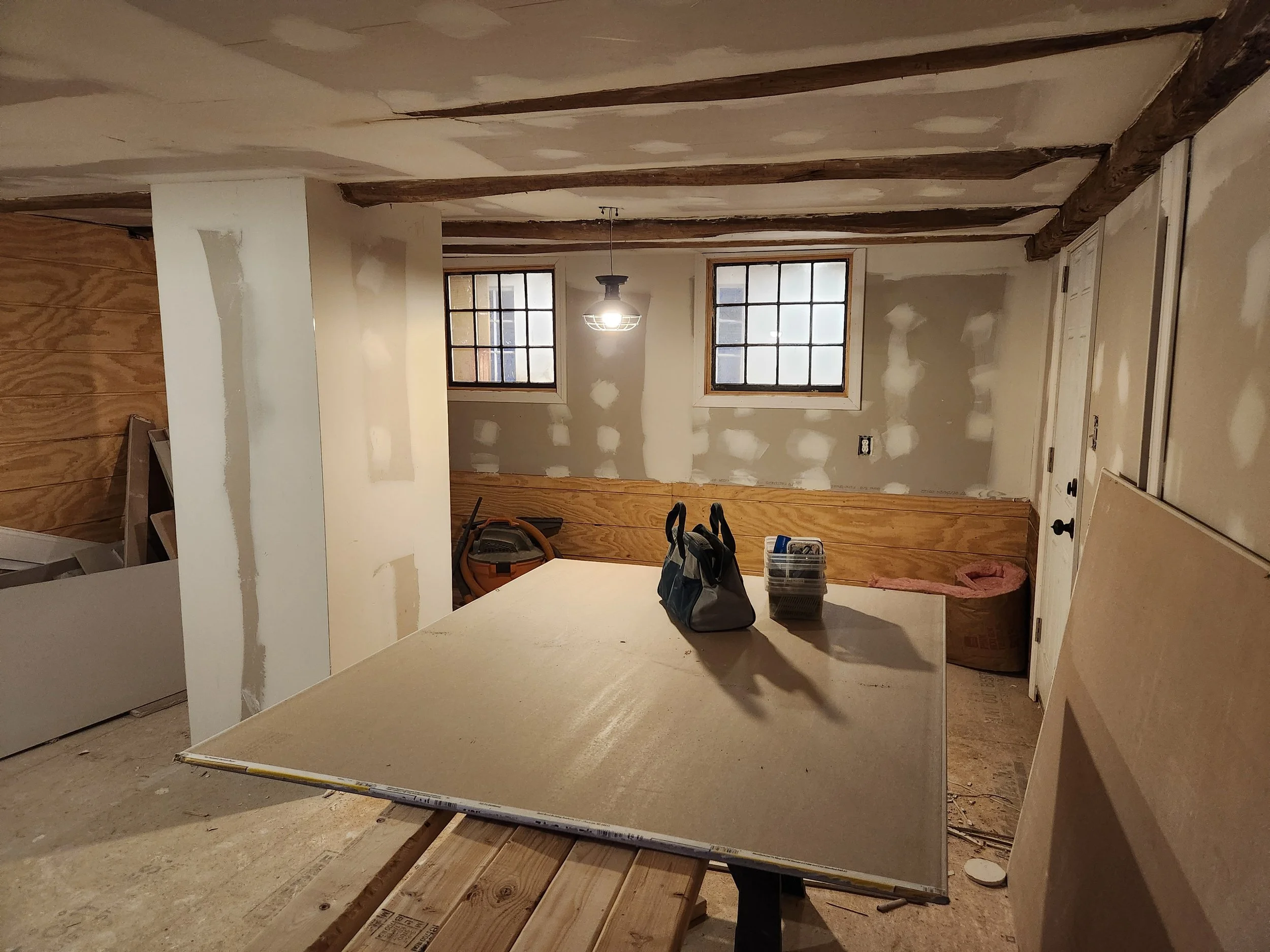 Interior of a room under construction with drywall and wood paneling, two windows, tools and supplies on a table, and exposed ceiling beams.