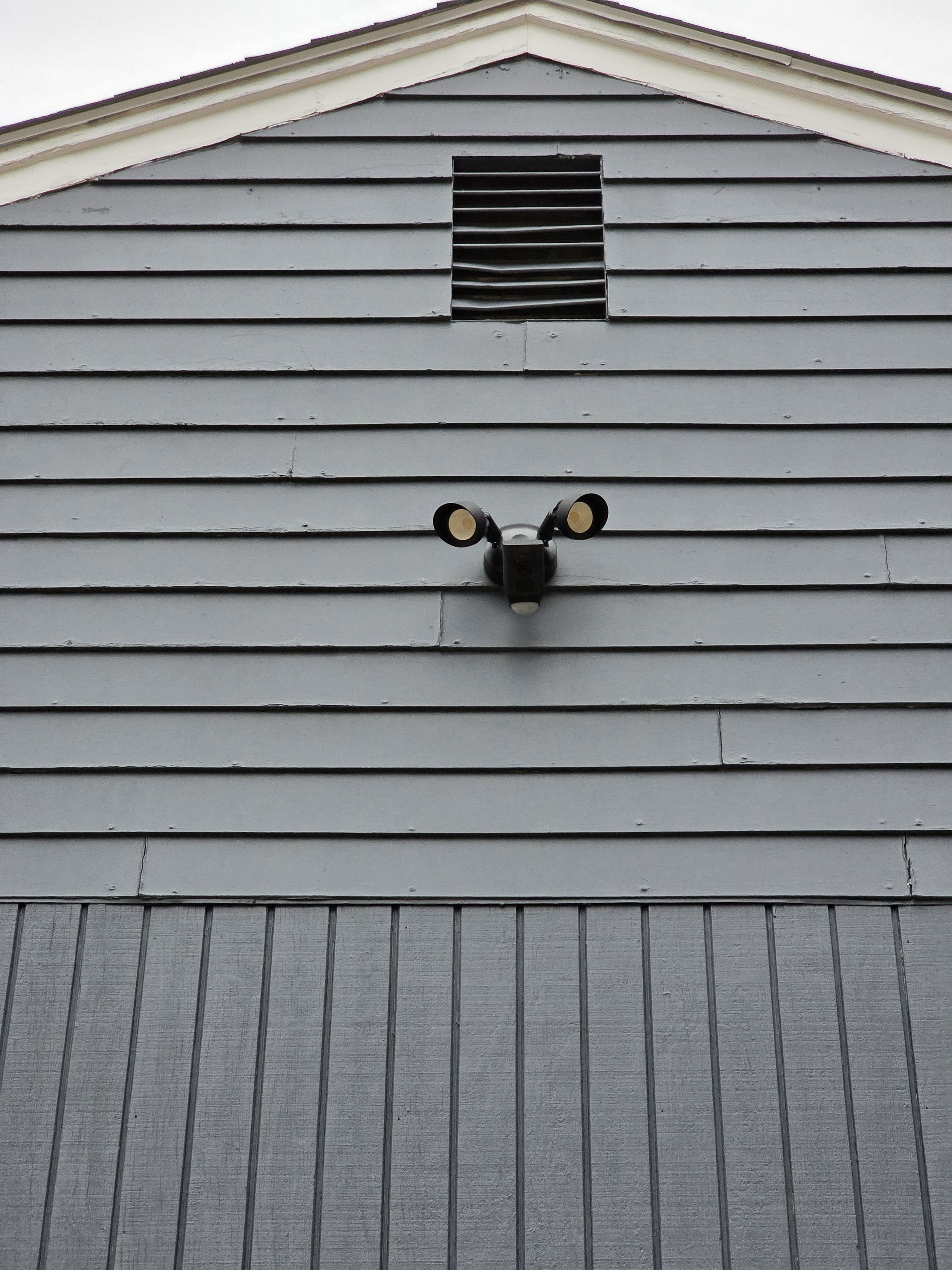Close-up of a building's exterior wall with light gray horizontal wooden siding, a black wall-mounted security camera, and a black rectangular vent near the roof.