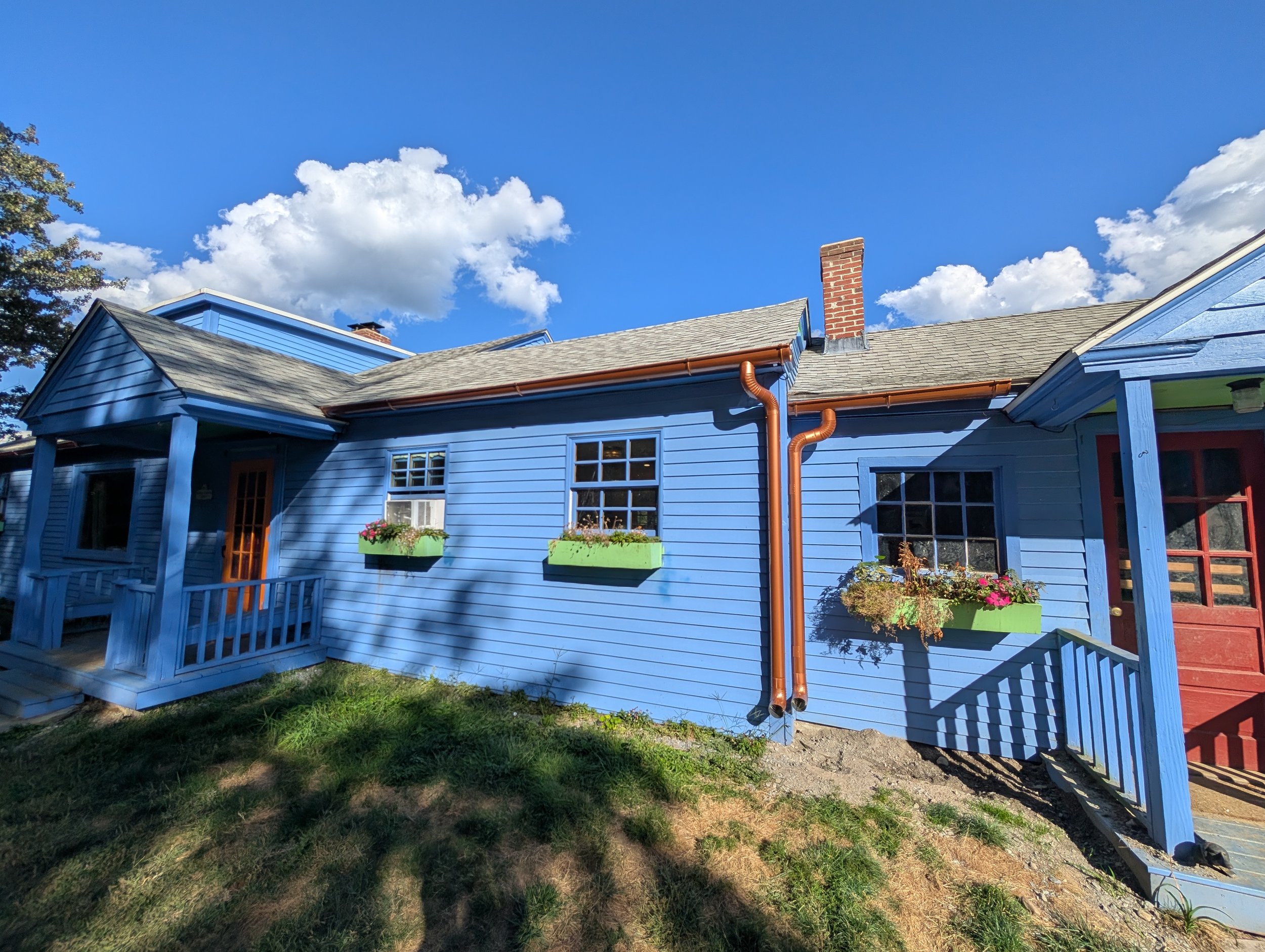 A bright blue house with flower boxes under the windows, a small porch with stairs, and a cloudy blue sky overhead.
