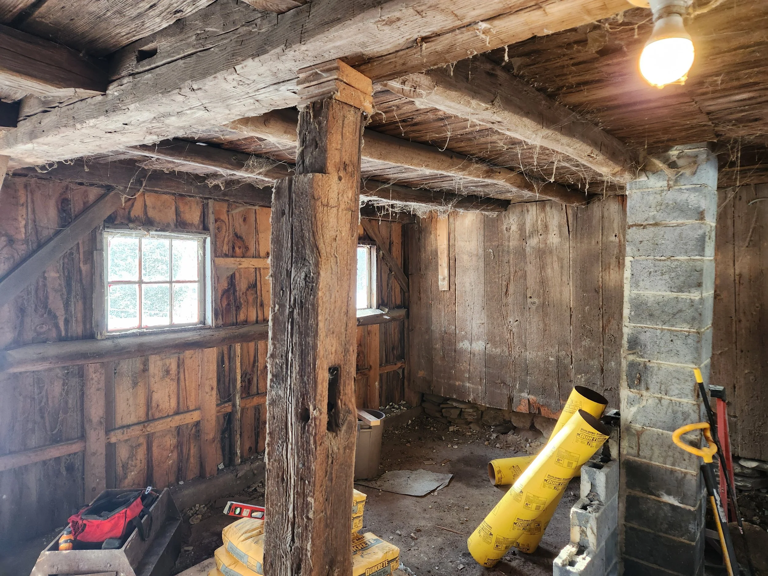 Interior of a rustic wooden barn with exposed beams, small windows, and construction supplies including yellow insulation rolls, a red toolbox, and various tools on the floor.