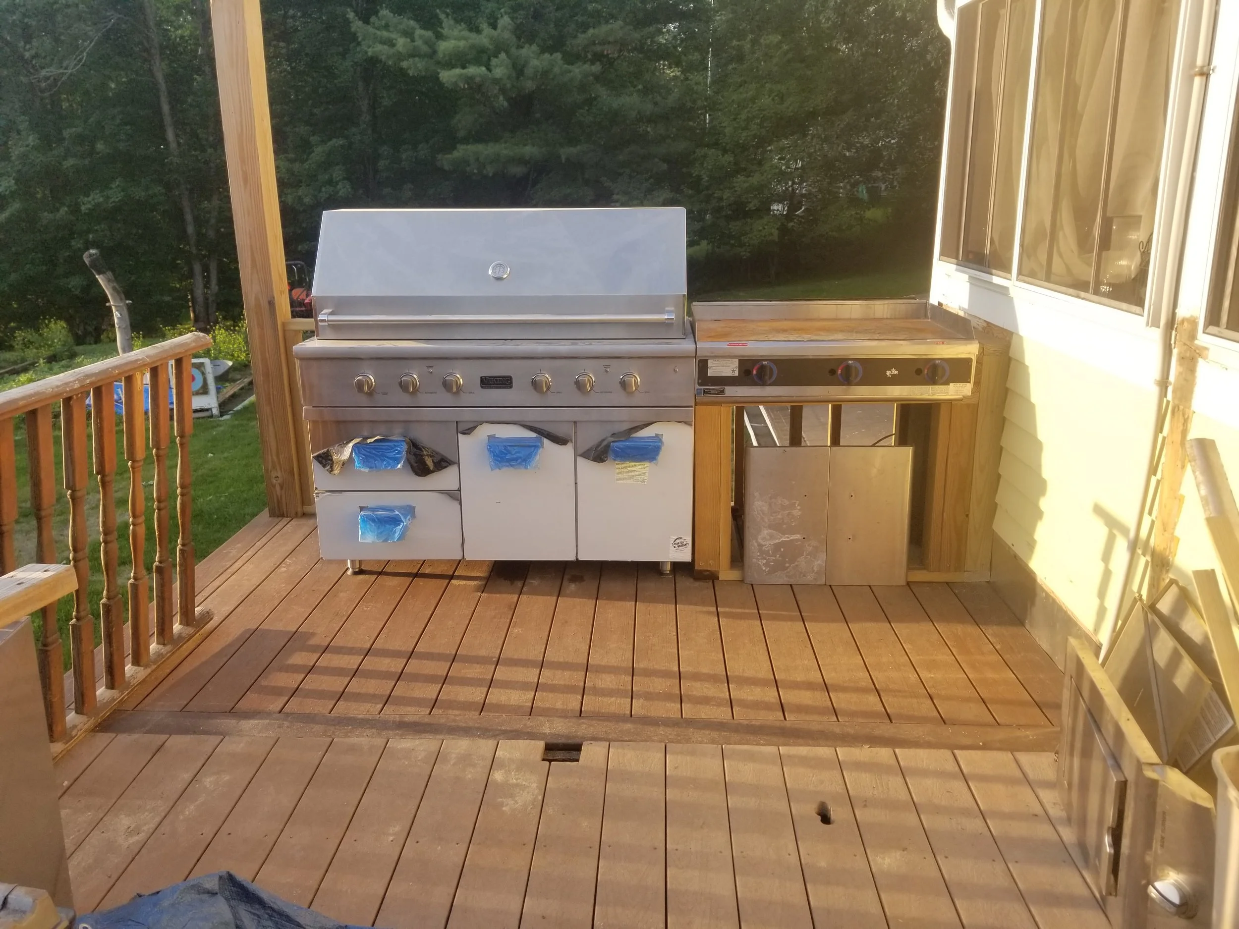 Unfinished outdoor kitchen on a wooden deck, featuring a stainless steel grill and a side burner with orange knobs, surrounded by a wooden railing, with a background of green trees and sunlight.