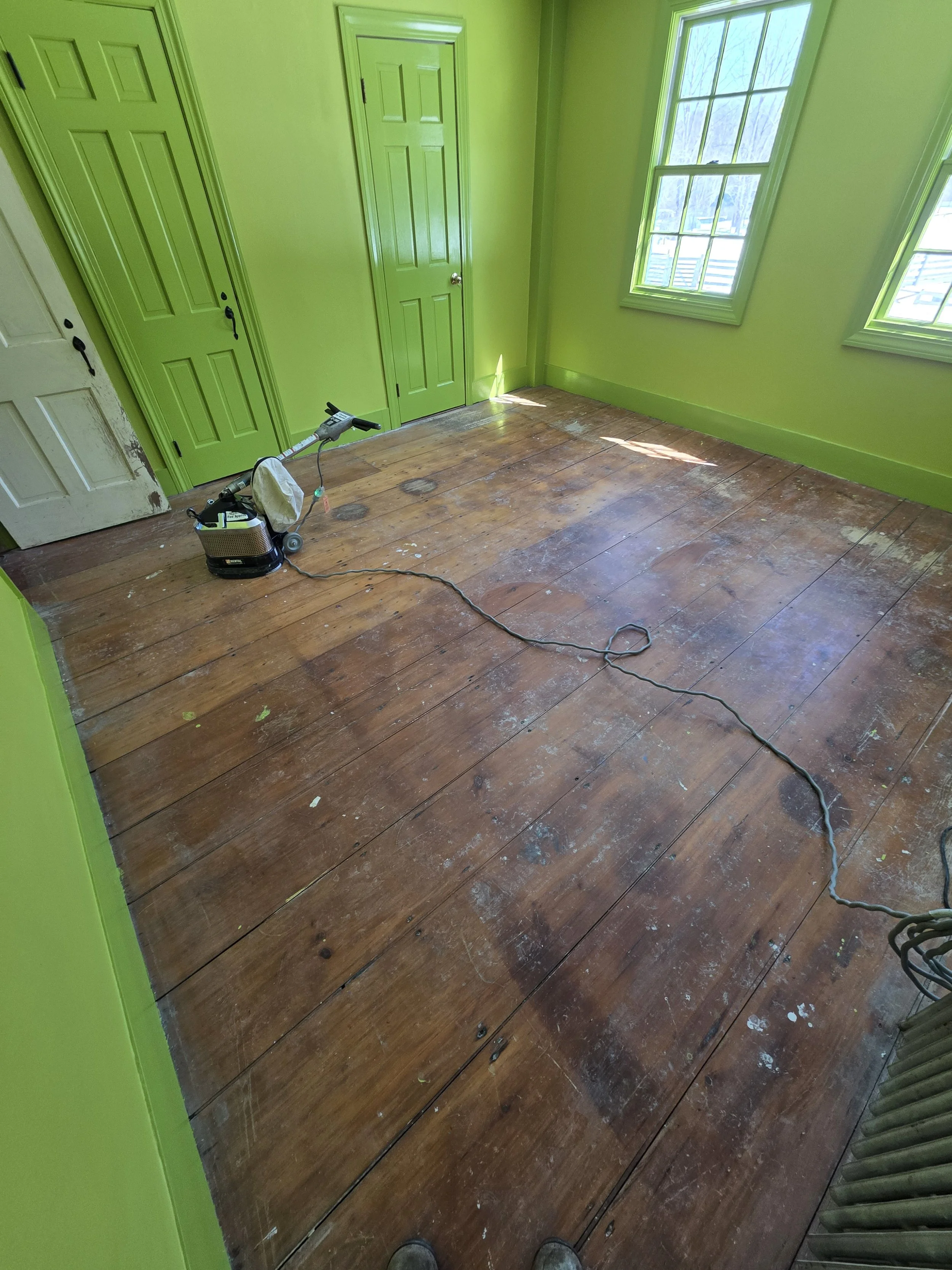 Empty room with bright green walls and trim, hardwood floor with paint splatters, and multiple windows letting in natural light.