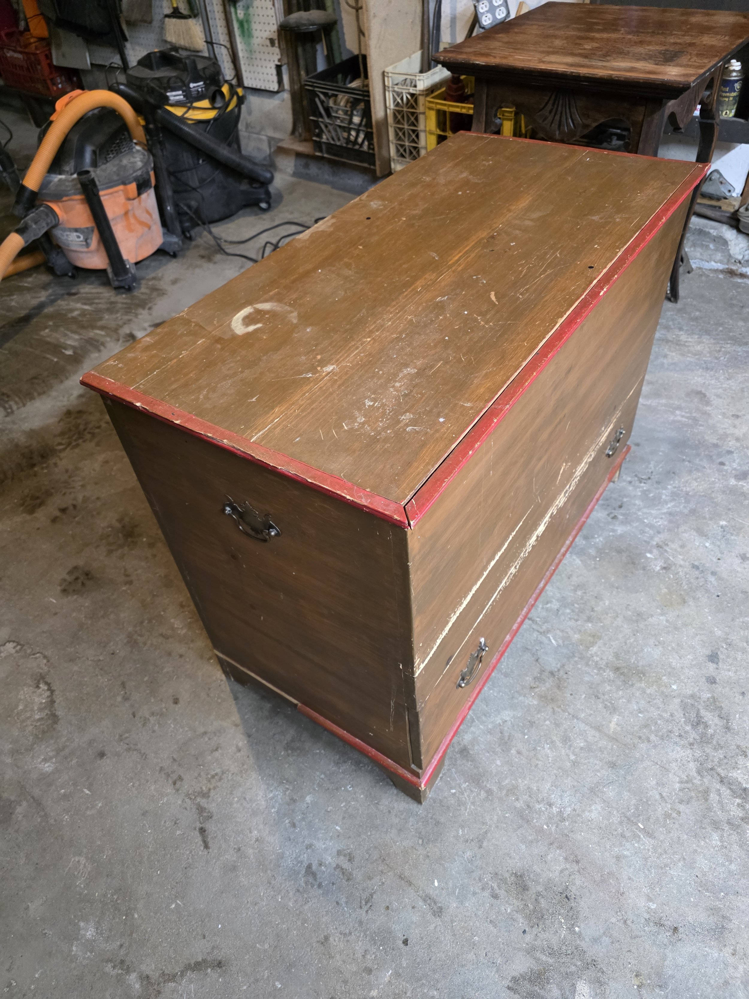 A wooden chest with metal handles and a red trim on the edges, placed on a concrete floor in a garage or workshop setting.
