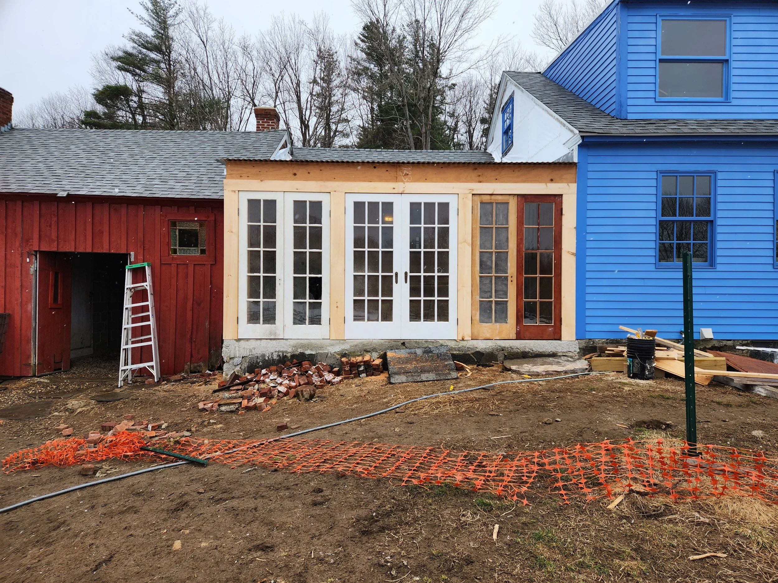 Construction site of a small extension with French doors attached to a red and blue house, a ladder leaning against the red part, and building materials scattered around.