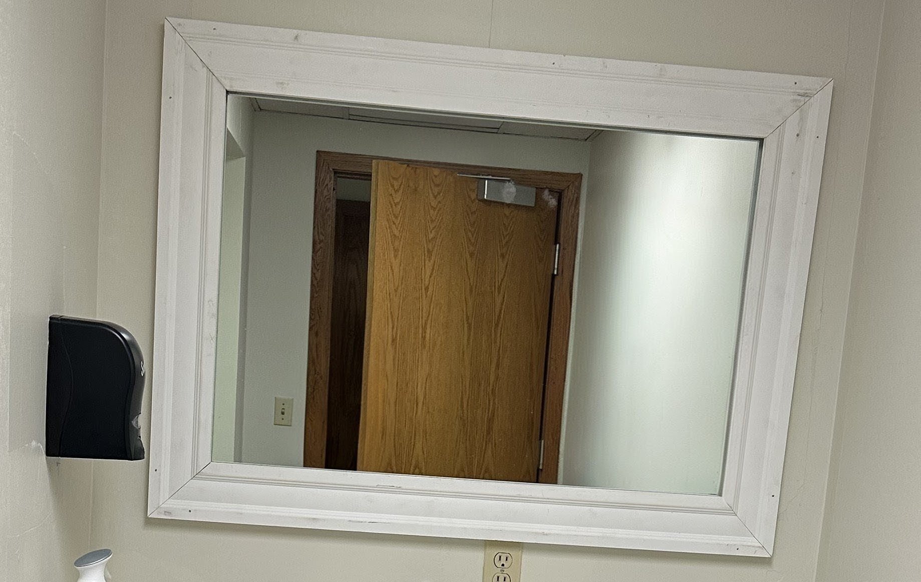 A large rectangular mirror with a white frame mounted on a beige wall, reflecting a wooden door and a beige room.