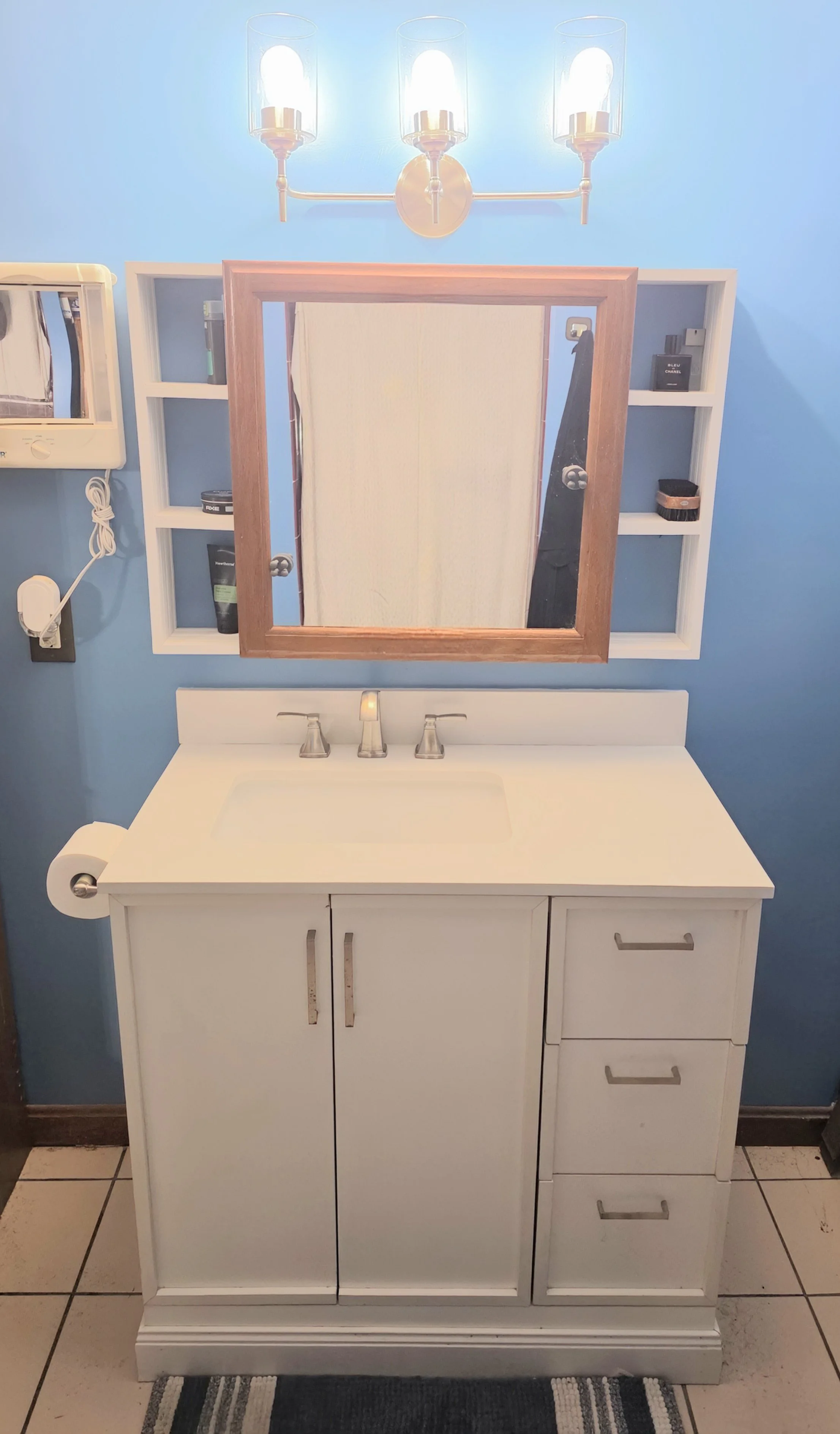 Bathroom vanity with a white cabinet, a mirror with a wooden frame, and a wall-mounted shelf with toiletries, against blue walls and tiled floor.
