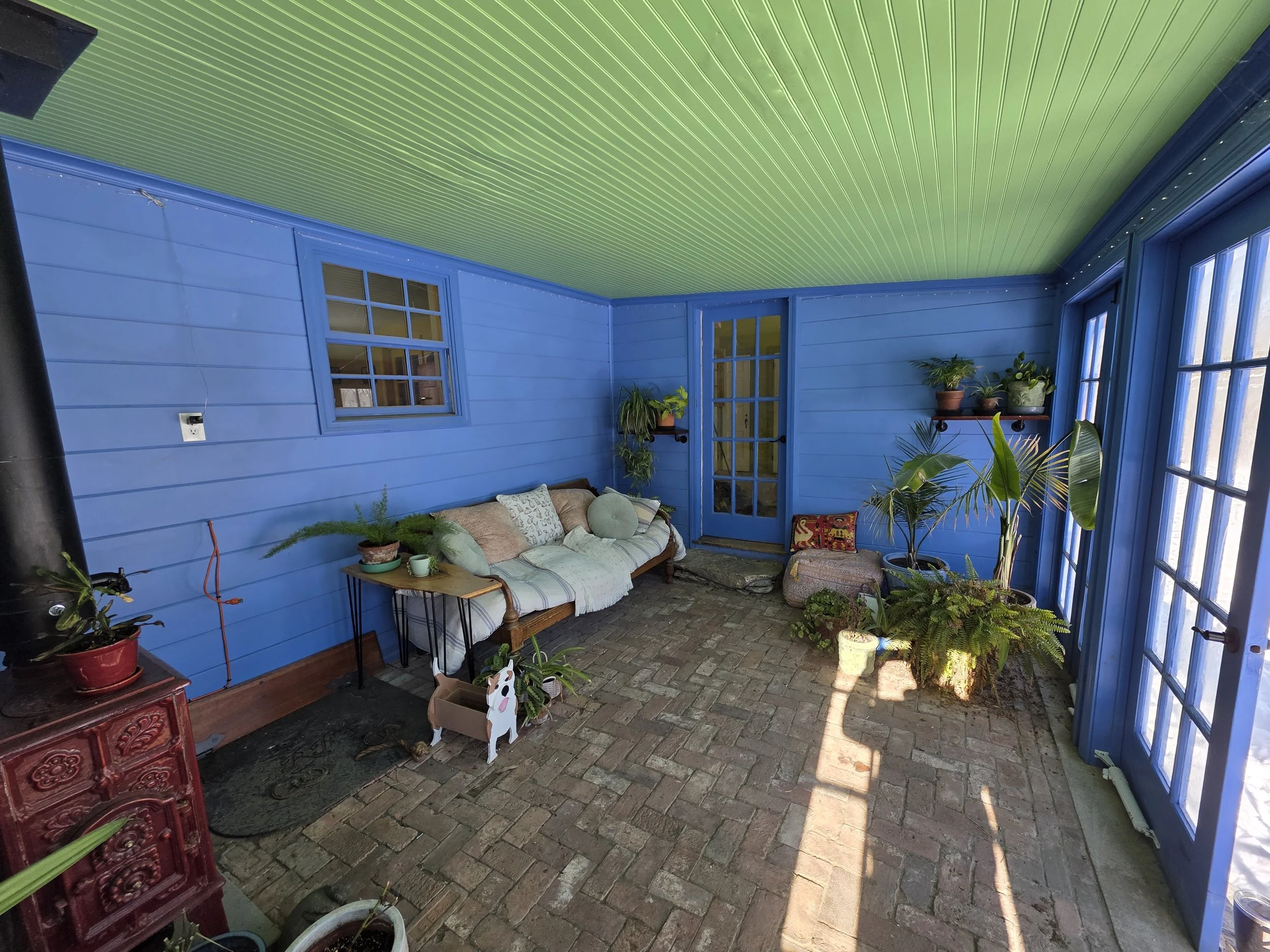 Enclosed porch with blue wooden walls, green shiplap ceiling, brick flooring, and large windows. Contains a wooden bench with pillows, houseplants on shelves and the floor, a small red stove, and door leading outside.