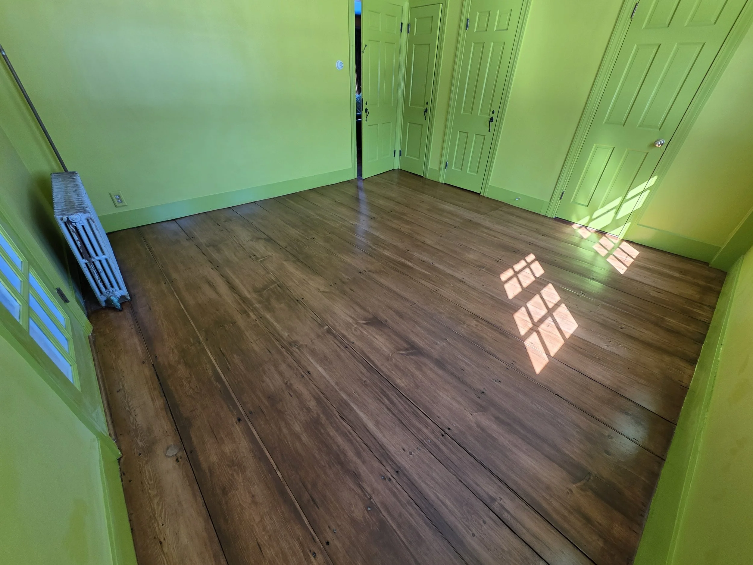 Empty room with bright green walls, wooden floor, closed green closet doors, and sunlight casting a shadow of a window on the floor.