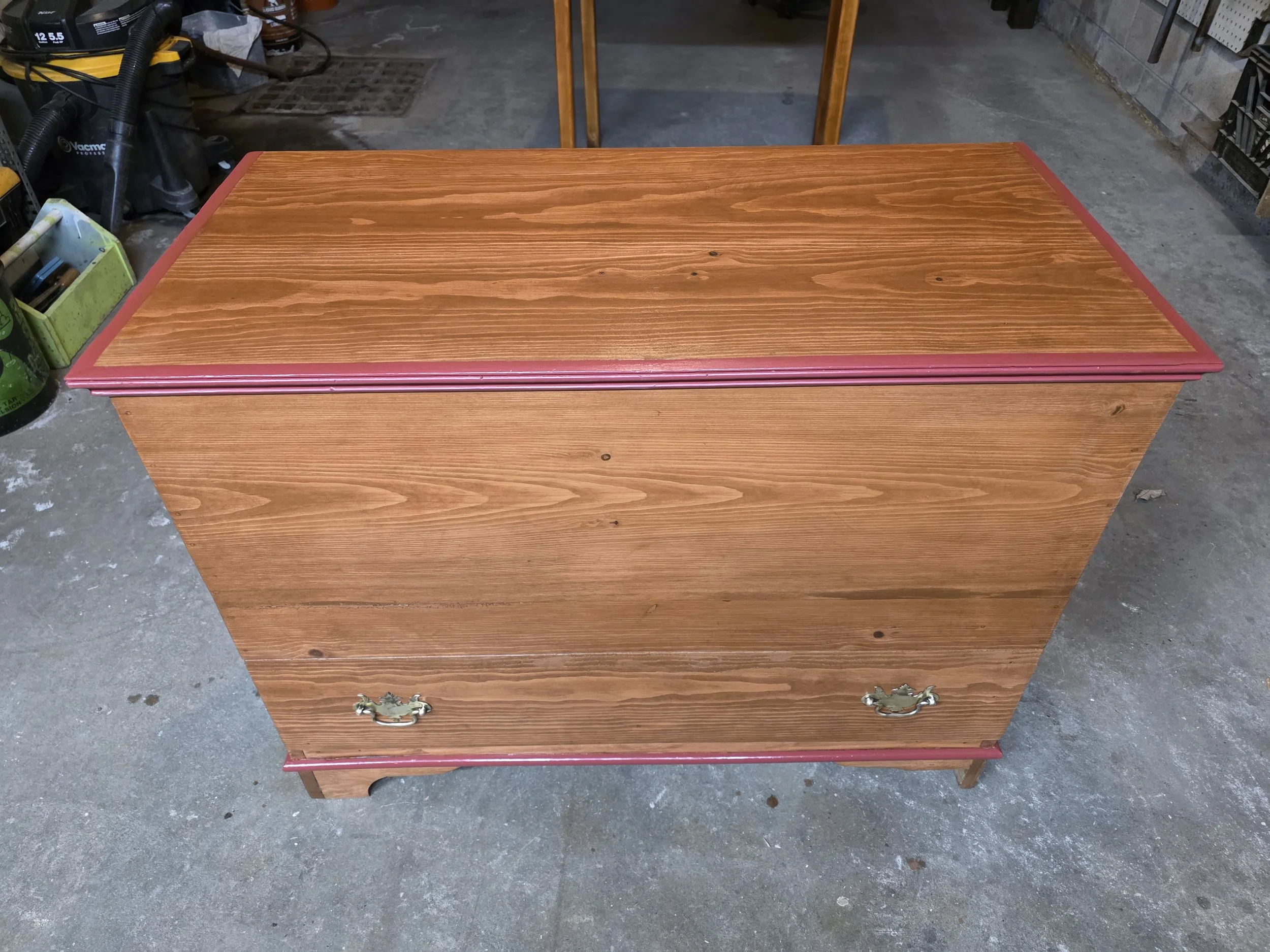 Wooden chest with brass handles and a pink trim on a concrete garage floor.