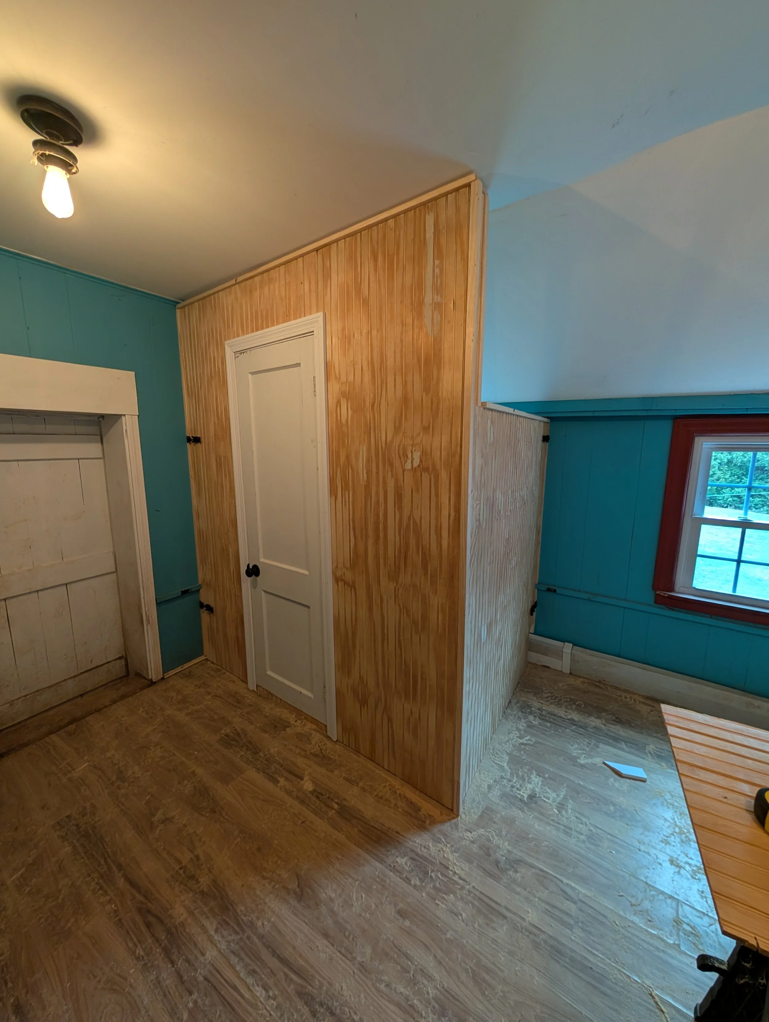 Interior room with wood flooring, a wood-paneled wall, a white door, and a window with red trim.