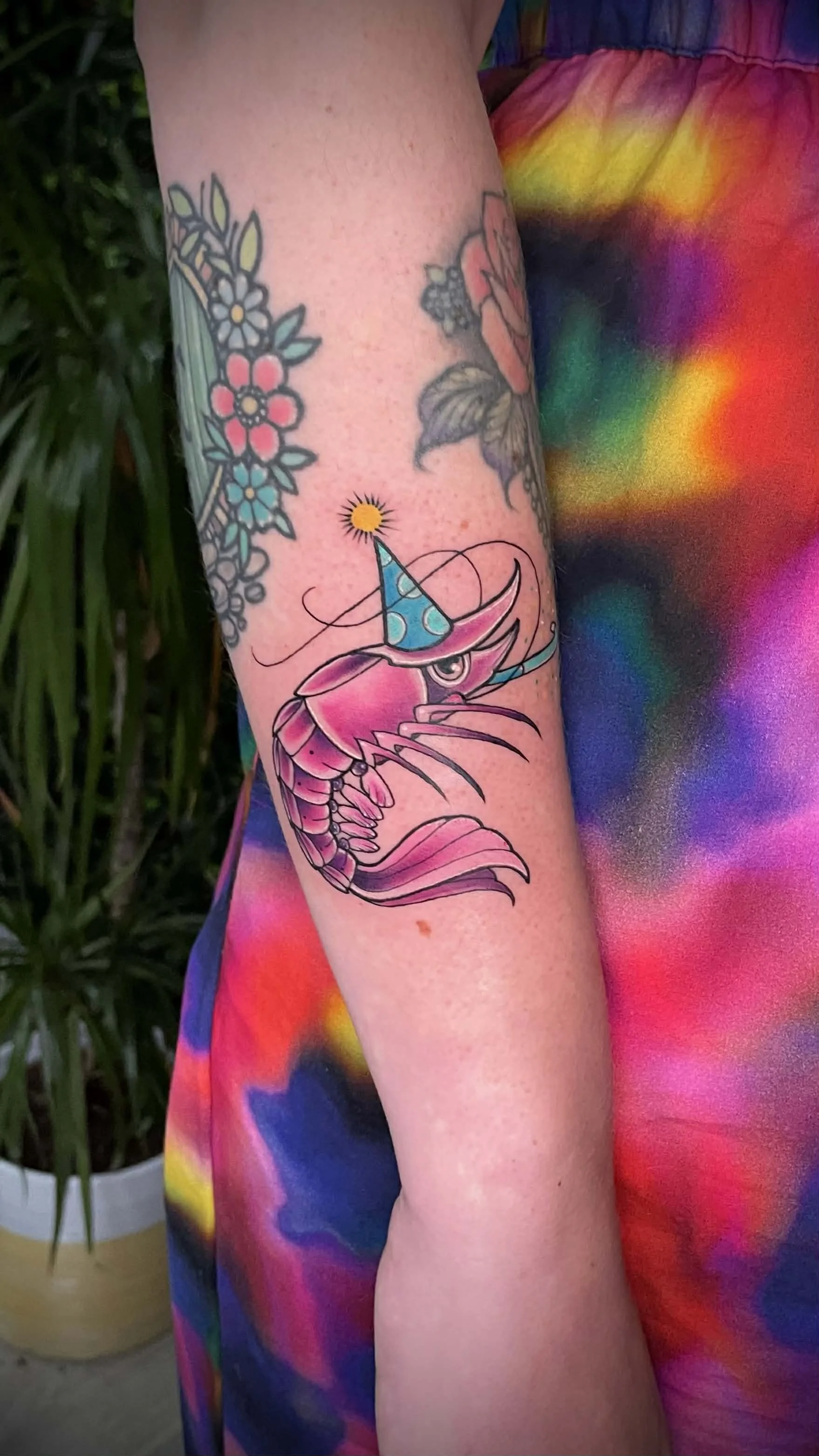 Colorful tattoo of a pink shrimp wearing a blue party hat with a yellow pom-pom and a rainbow-colored scarf, on a person's arm.