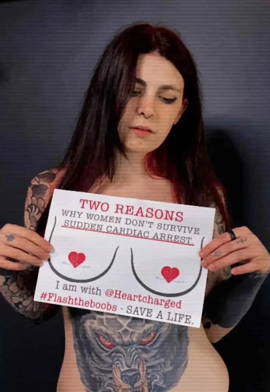 Woman with dark hair and tattoos holding a sign that discusses women's health, featuring a graphic of a woman's chest with a heart and text emphasizing reasons women don't survive sudden cardiac arrest.