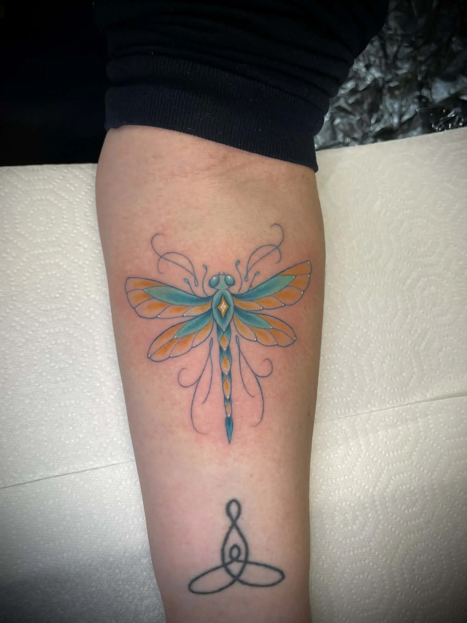 Colorful tattoo of a dragonfly with orange and teal wings on a person's upper arm, with another simpler black ink tattoo below it.