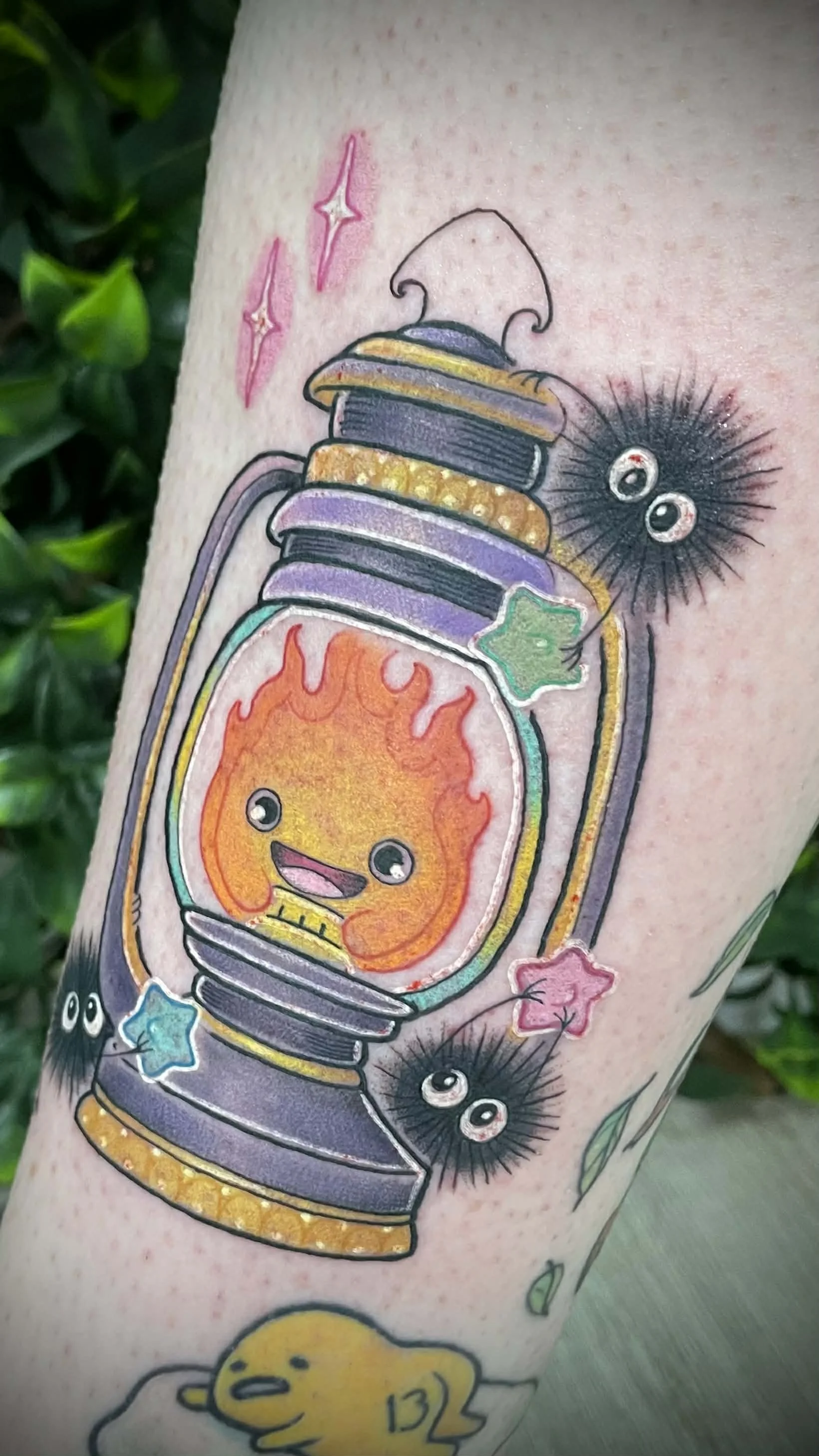Colorful tattoo of a smiling flame with big eyes inside a vintage lantern, surrounded by small cheerful cartoon-like black fuzzy creatures with big eyes, and pink, blue, and green star-shaped accents. Howls moving castle studio ghibli