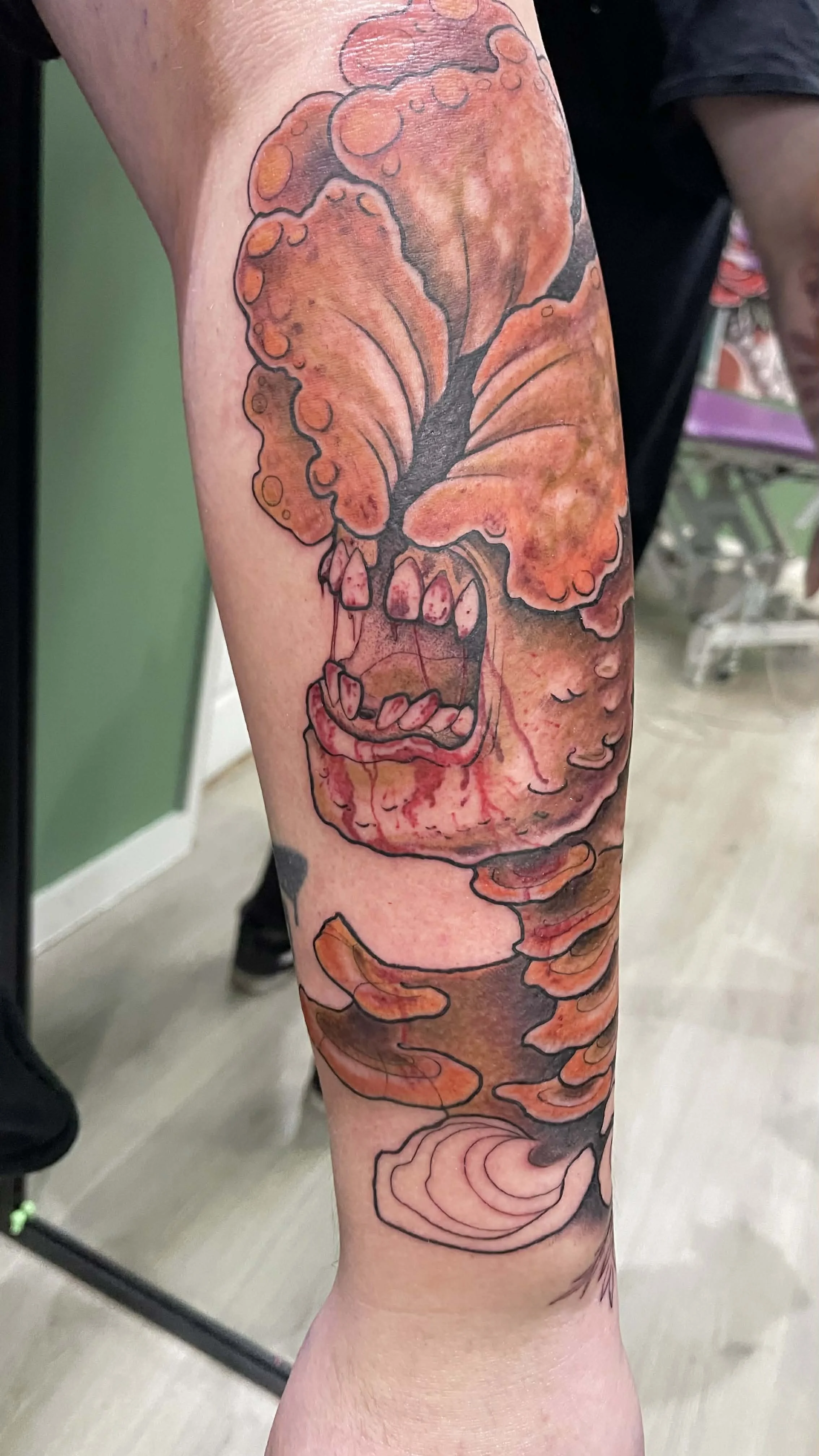 A tattoo of a smoking, angry mushroom with a mouth and teeth, in orange and pink hues, with black outlines. The mushroom appears to be crying or oozing in red.the last of us clicker tattoo 