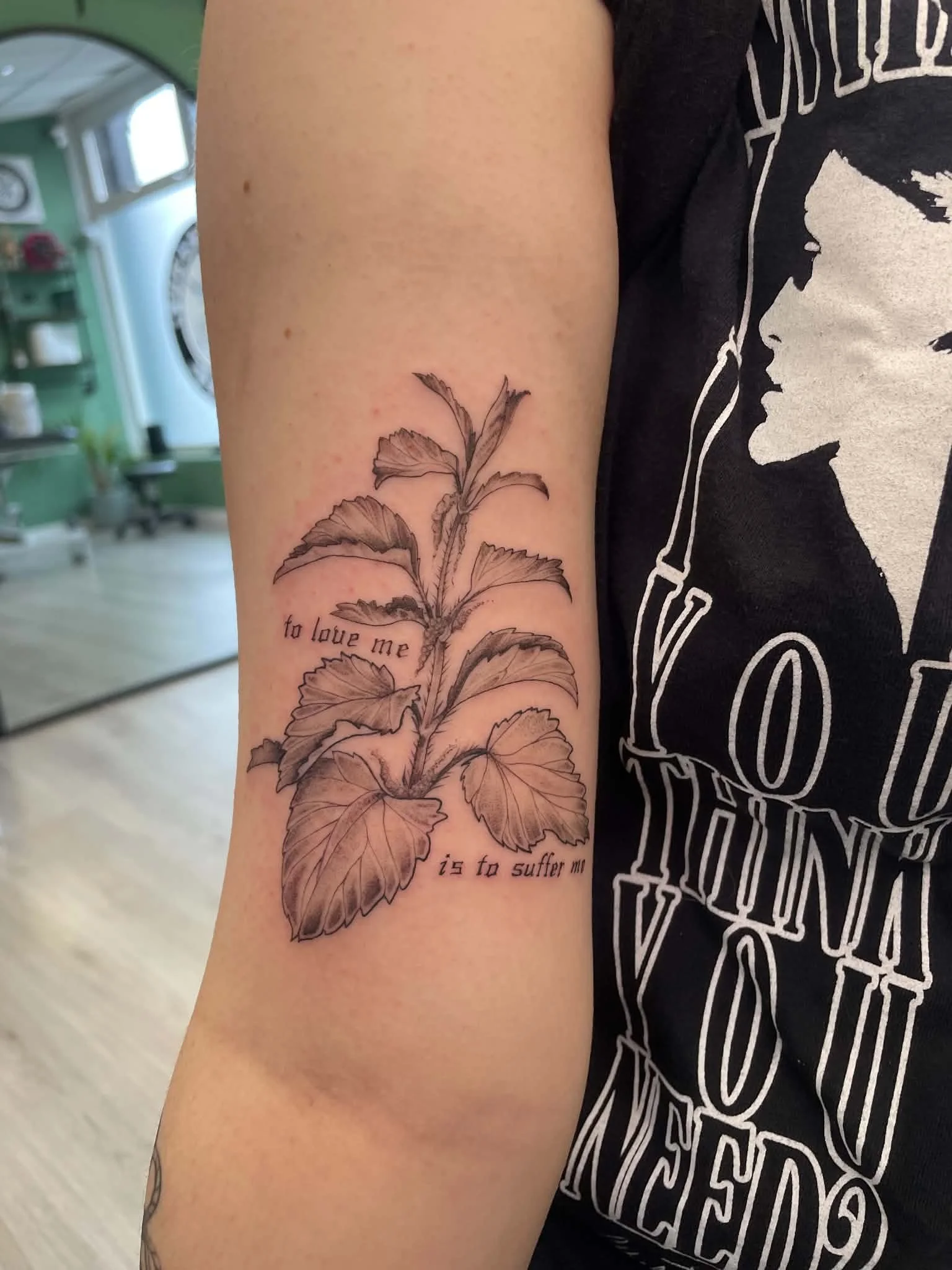 Tattoo of a plant with leaves on a person's upper arm, with partially visible text underneath the plant. The background shows an indoor setting with a washing machine and shelves.