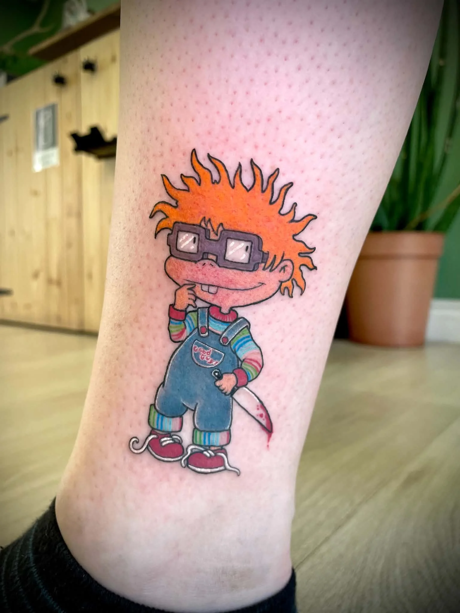 Colorful tattoo of a cartoon character with orange, spiky hair, wearing glasses, striped shirt, overalls, and sneakers, on a person's thigh. Chuckle chucky