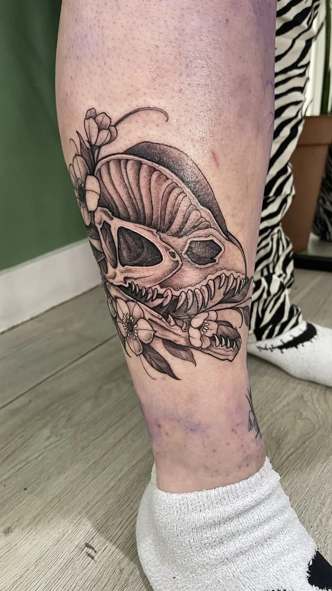 Black and gray tattoo of a dinosaur skull with flowers and plants around it on a person's leg.