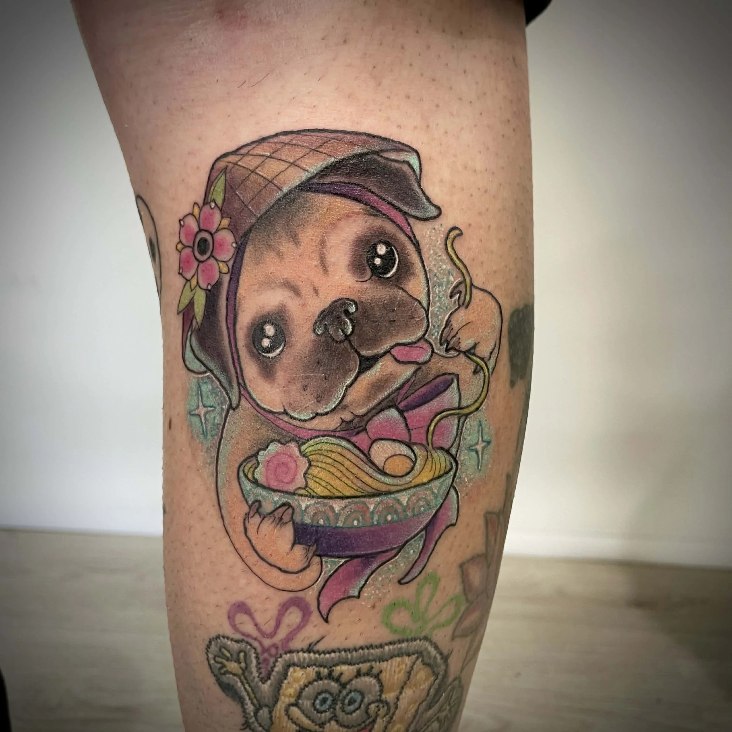 Tattoo of an adorable puppy with big eyes, wearing a bonnet with a flower, holding a bowl of ramen with soup and noodles, surrounded by decorative stars and hearts.