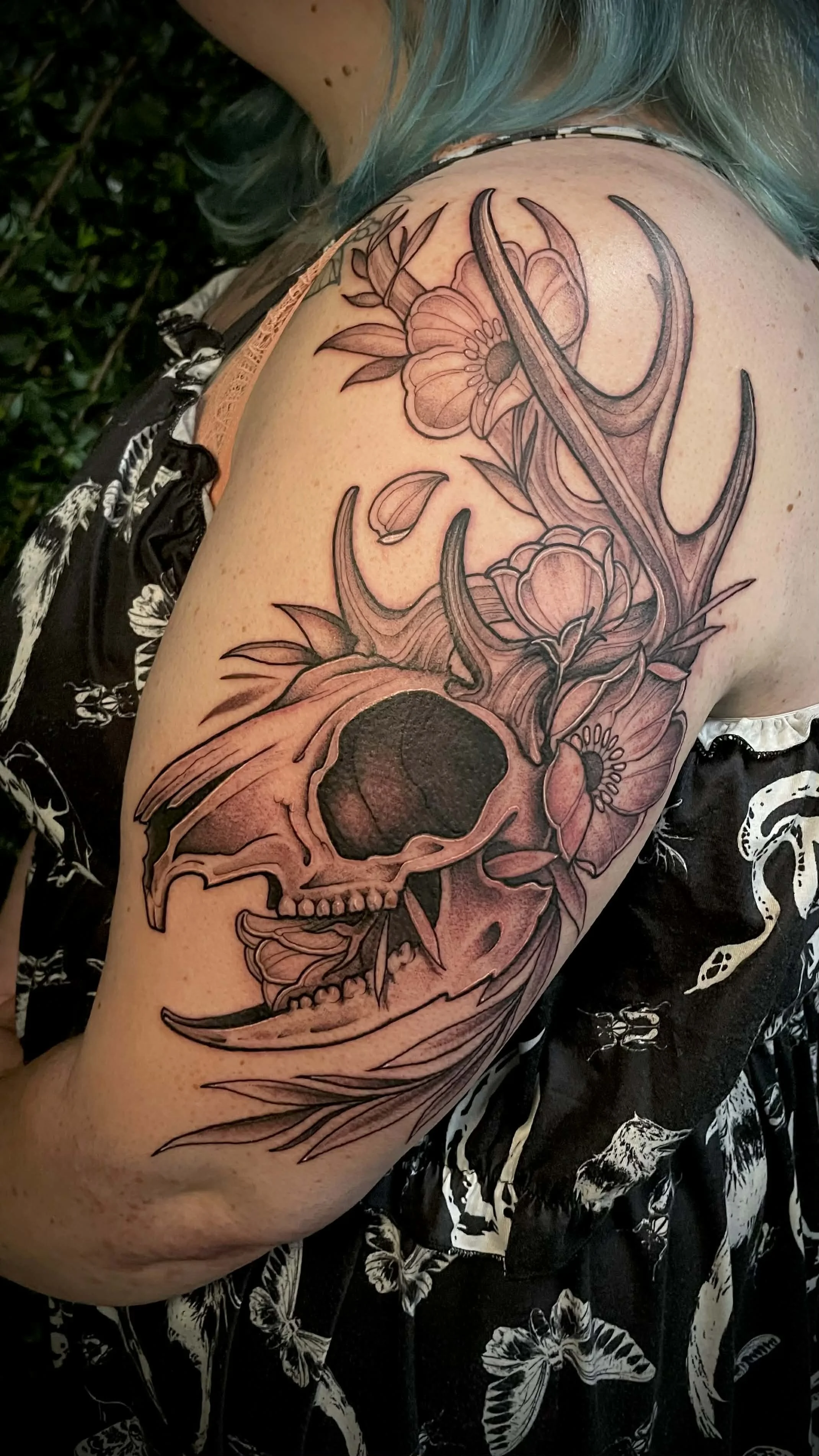 Tattoo of a jackalope skull with antlers, surrounded by flowers and leaves, on a person's upper arm and shoulder.