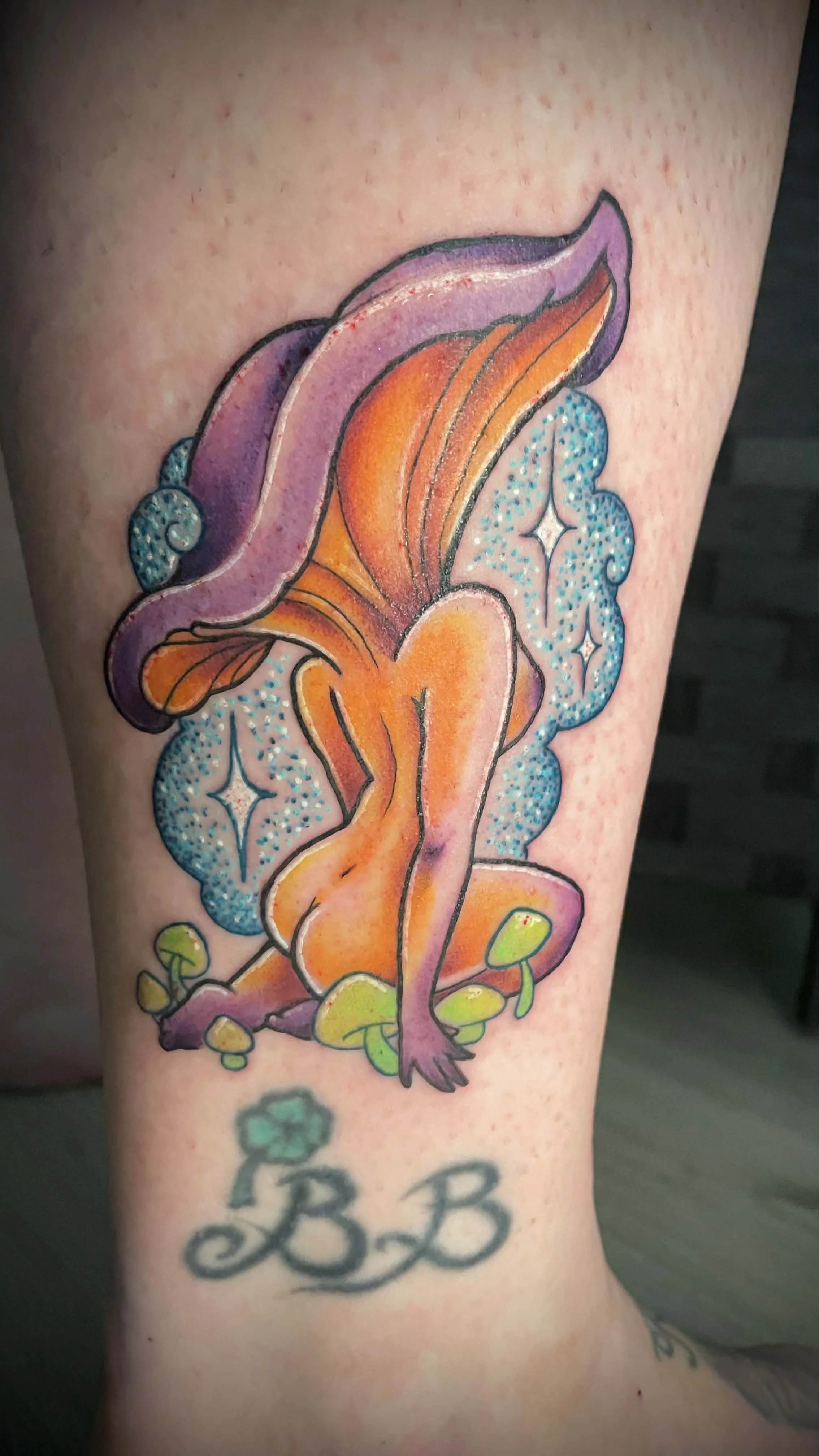 Colorful tattoo of a fairy girl with purple hair, orange wings, and a blue sparkly background, with initials 'B B' and a small green shamrock at the bottom.