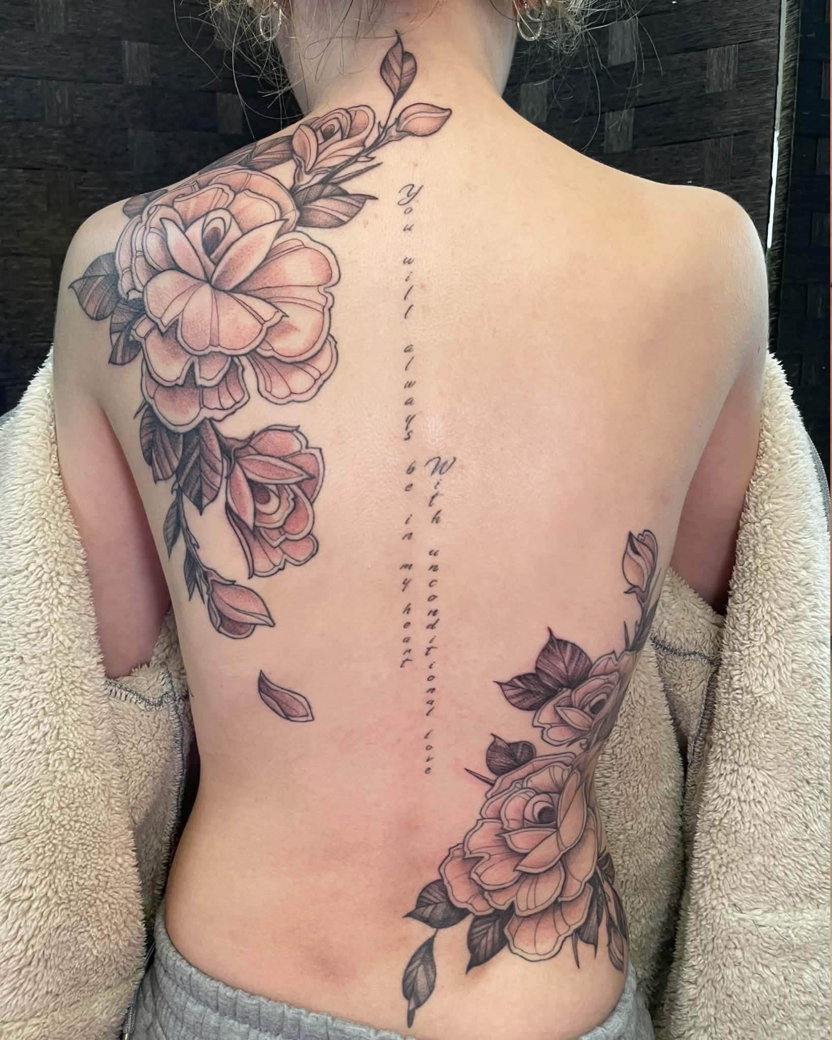 Tattoo of pink flowers with dark leaves and stems on the shoulder and back, with a vertical line of cursive script running down the center of the back.