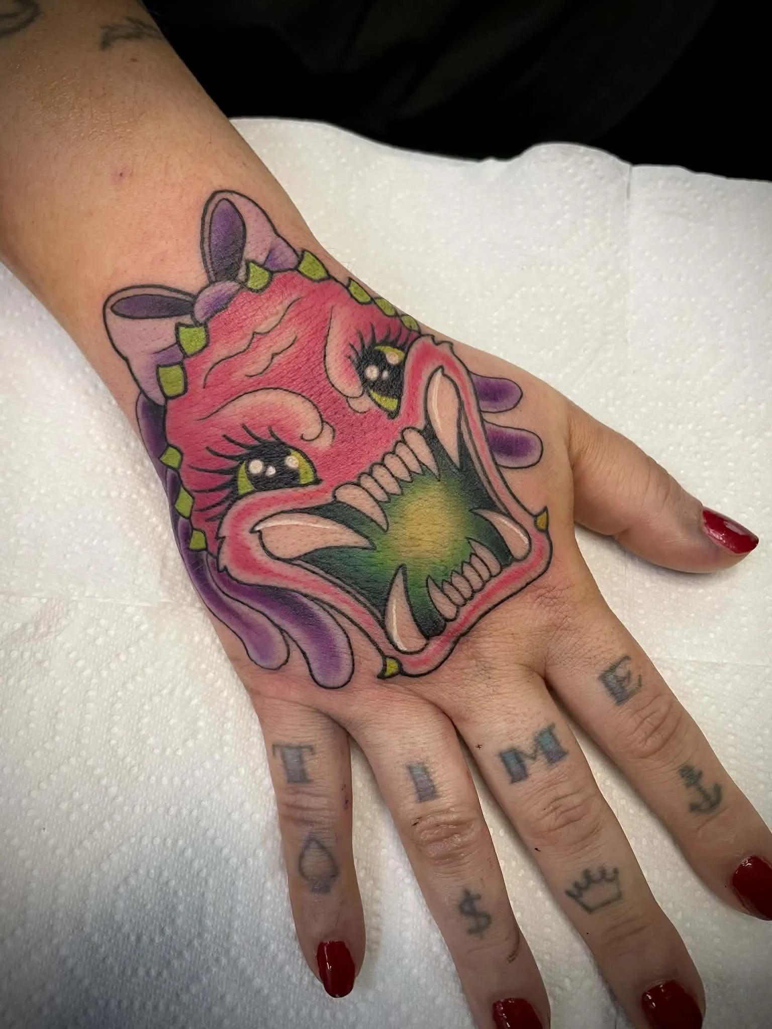 Tattoo of a cartoonish pink predator with large yellow eyes and an open mouth, showing sharp teeth, surrounded by purple and green accents on a person's hand.
