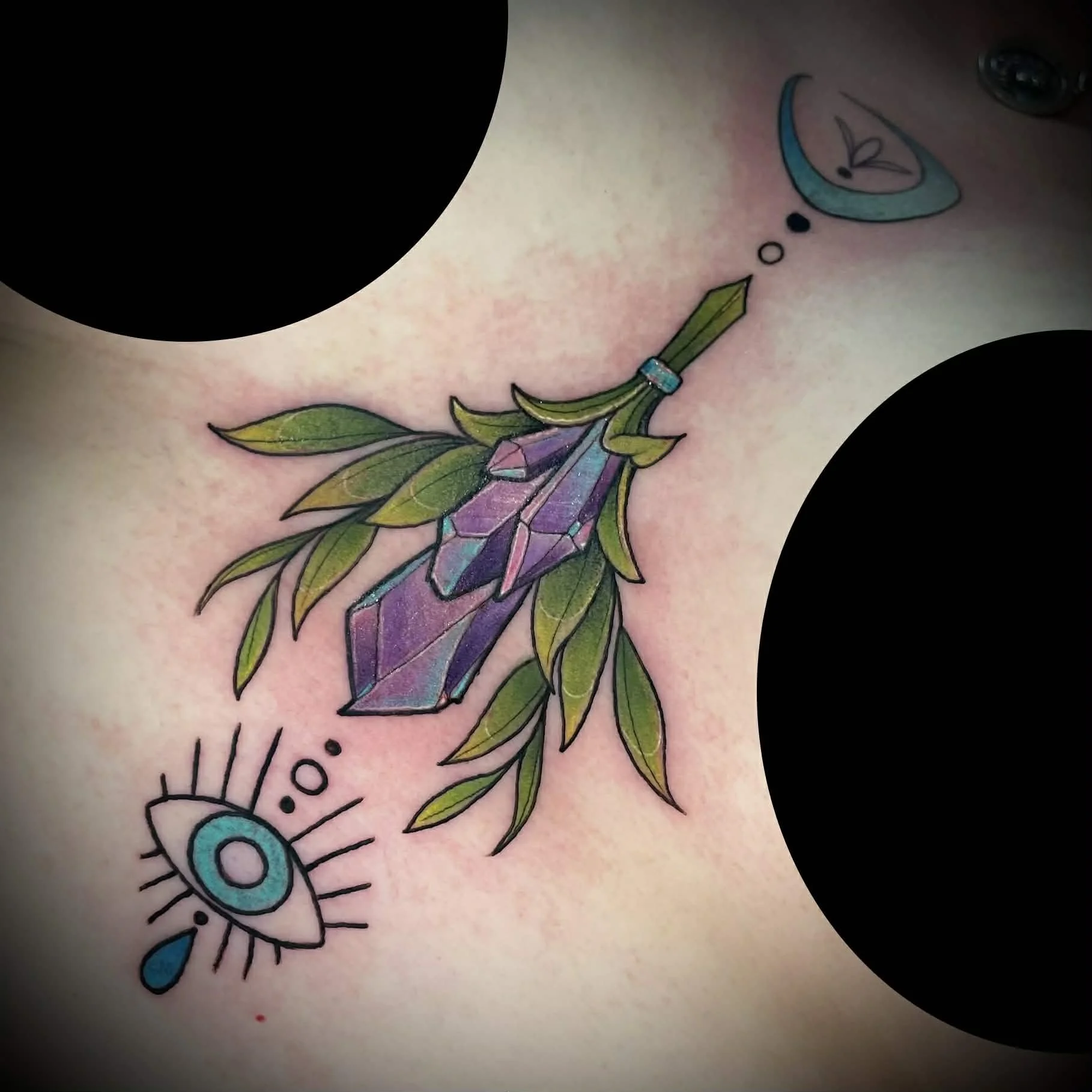 Colorful tattoo of a crystal pendant with green leaves, and a small eye with a teardrop below it on skin.