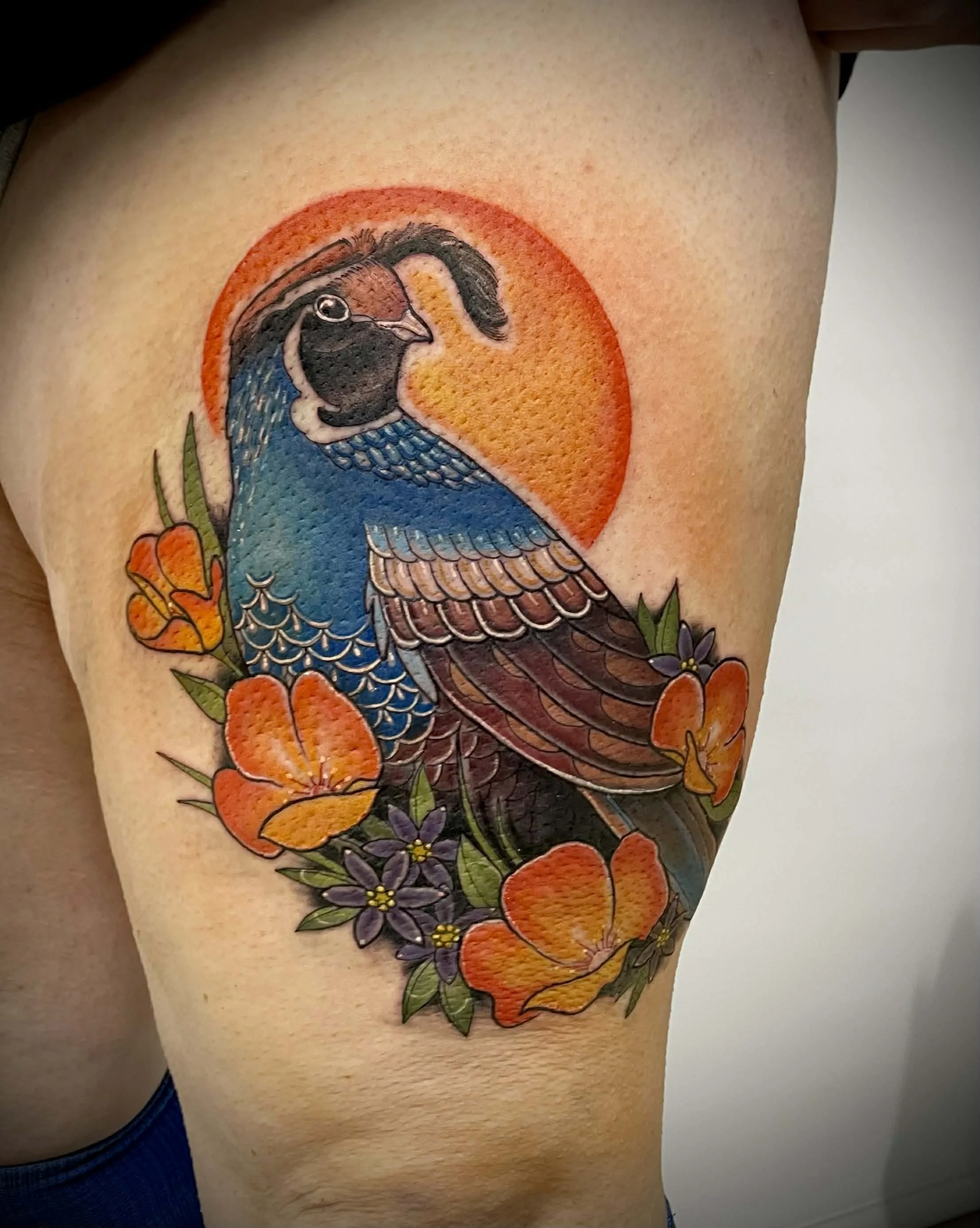 Colorful tattoo of a bird with a blue body and brown wings, surrounded by orange and purple flowers, with an orange sun in the background.
