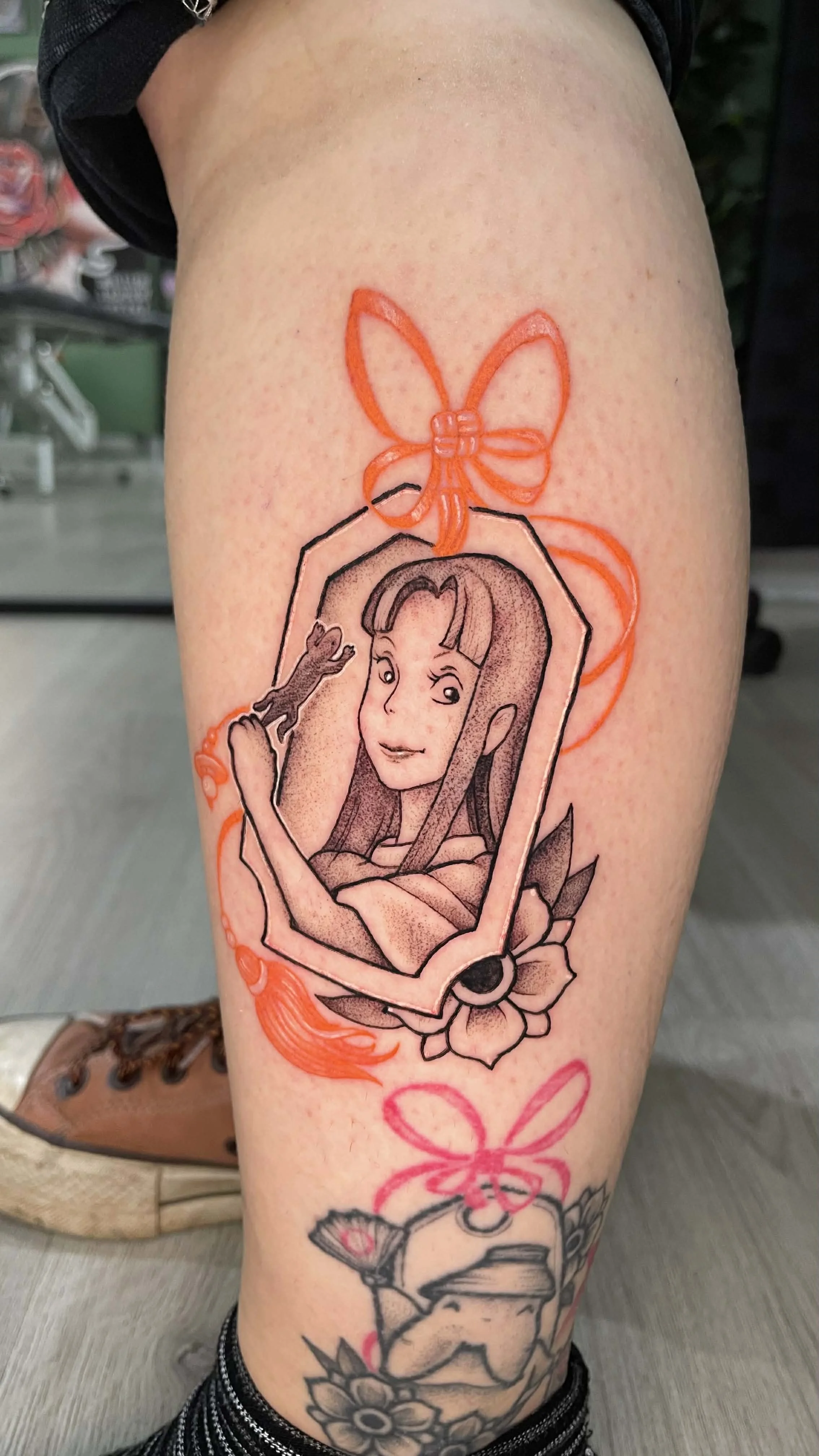 Tattoo on person's thigh featuring a stylized portrait of a woman with a frog on her hand, framed within an outline, decorated with a flower and an orange bow.