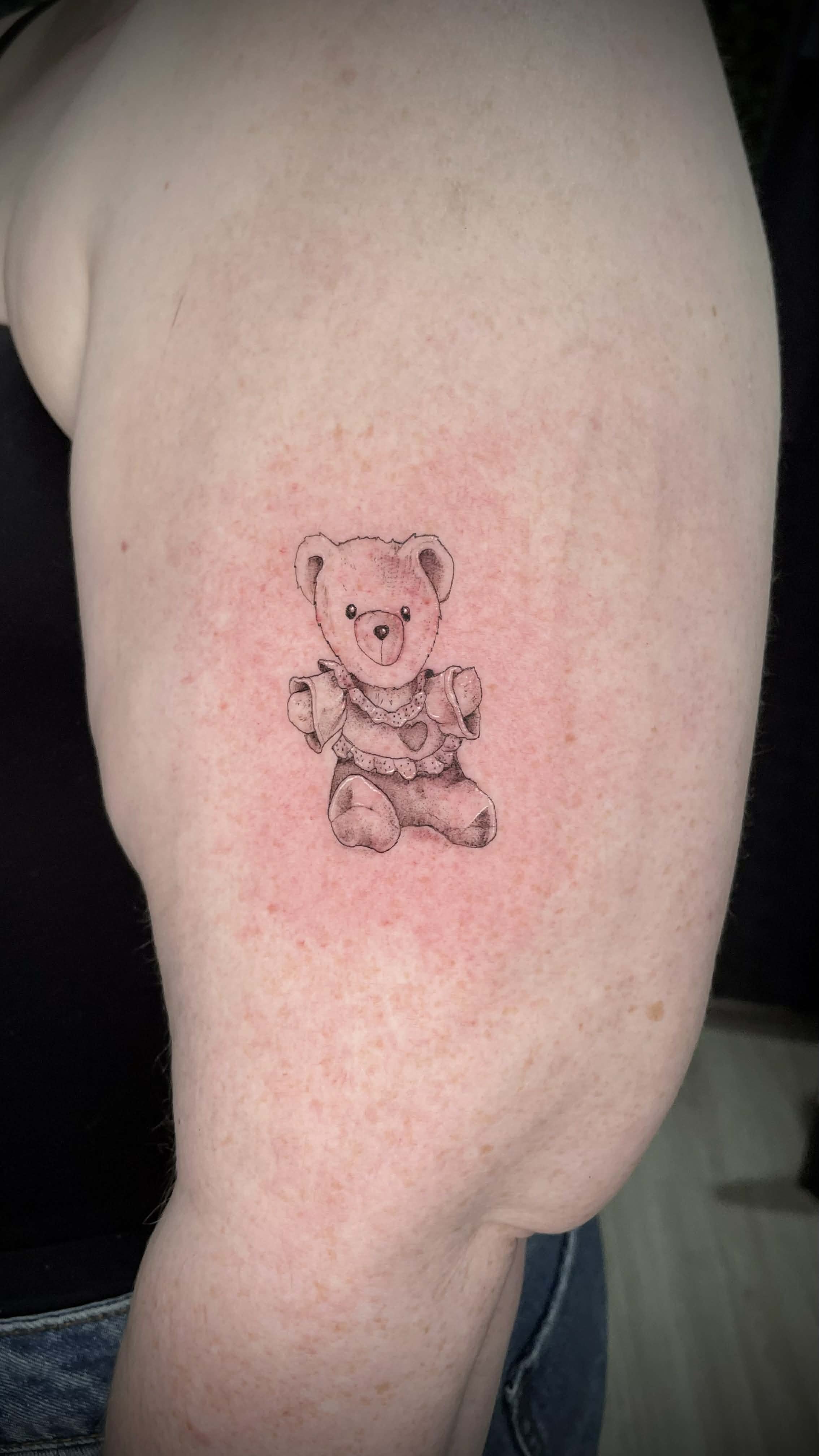 Tattoo of a cute teddy bear in a dress on person's upper arm.