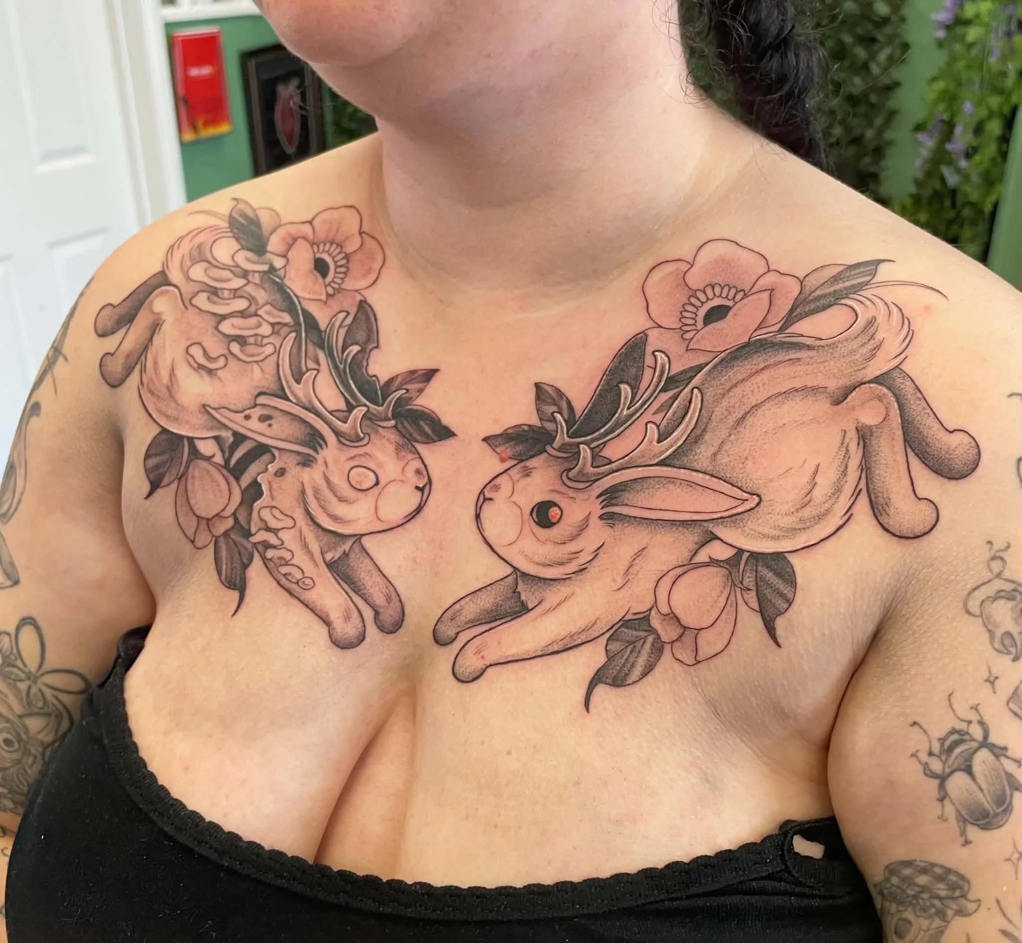 Close-up of a person's shoulder and chest with an intricate black and gray floral tattoo. The tattoo features roses, leaves, and a rabbit, and is detailed and shaded. The person is wearing a black top with a lace edge, and part of their black hair is