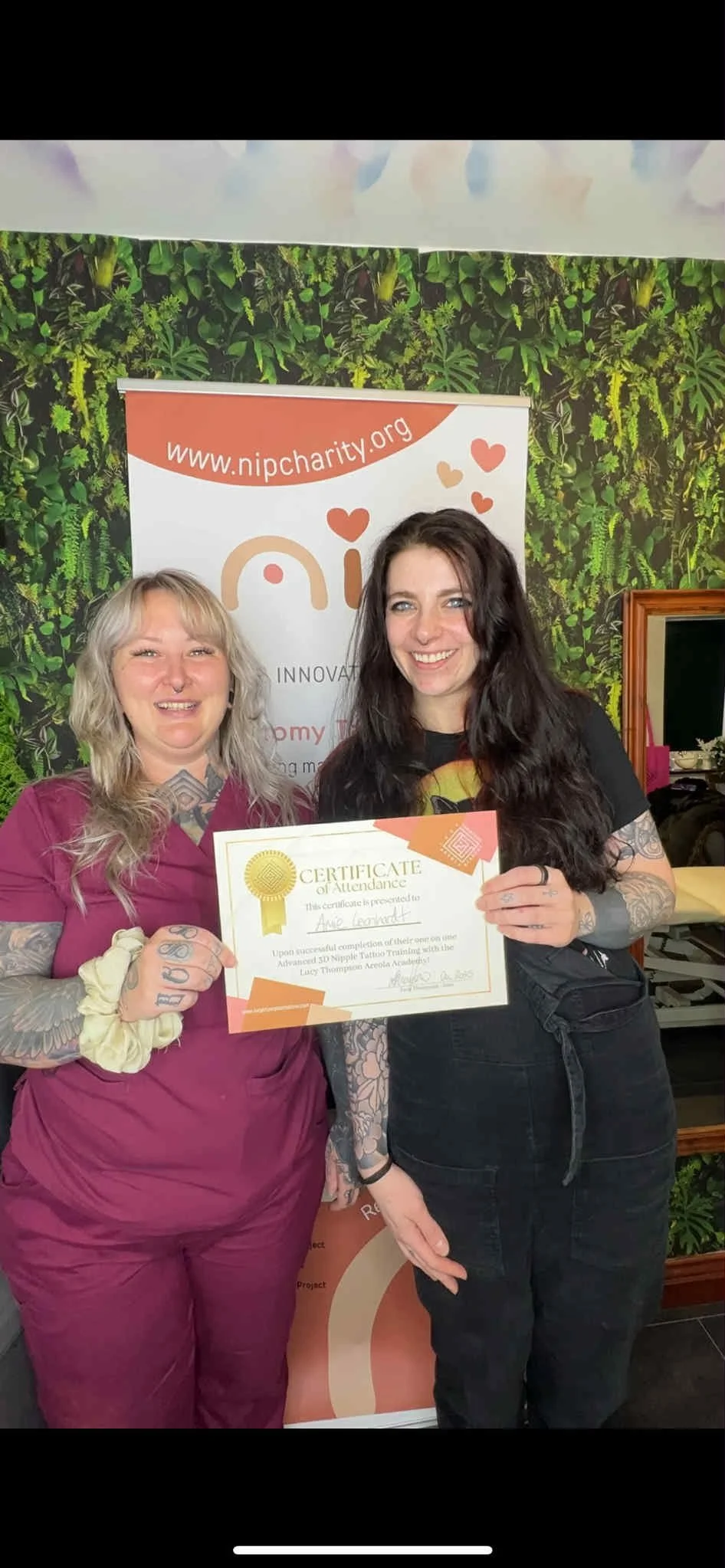 Two women standing together holding a certificate of attendance at a tattoo training workshop, with a backdrop displaying the website www.nipcharity.org and a leafy green wall.