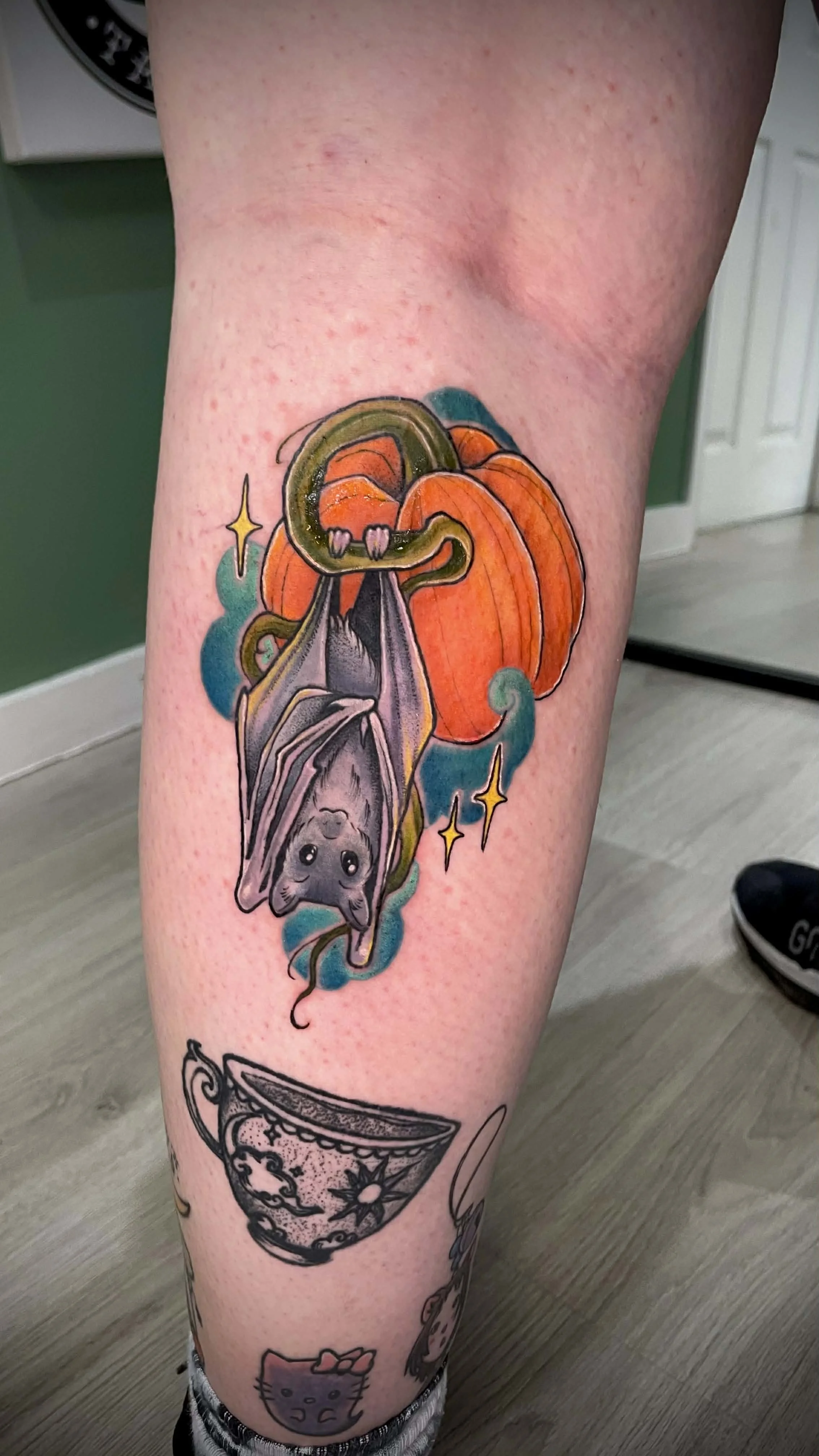 Tattoo of a cute gray bat hanging upside down with a smiling expression, set against an orange pumpkin, with sparkles and a blue background.