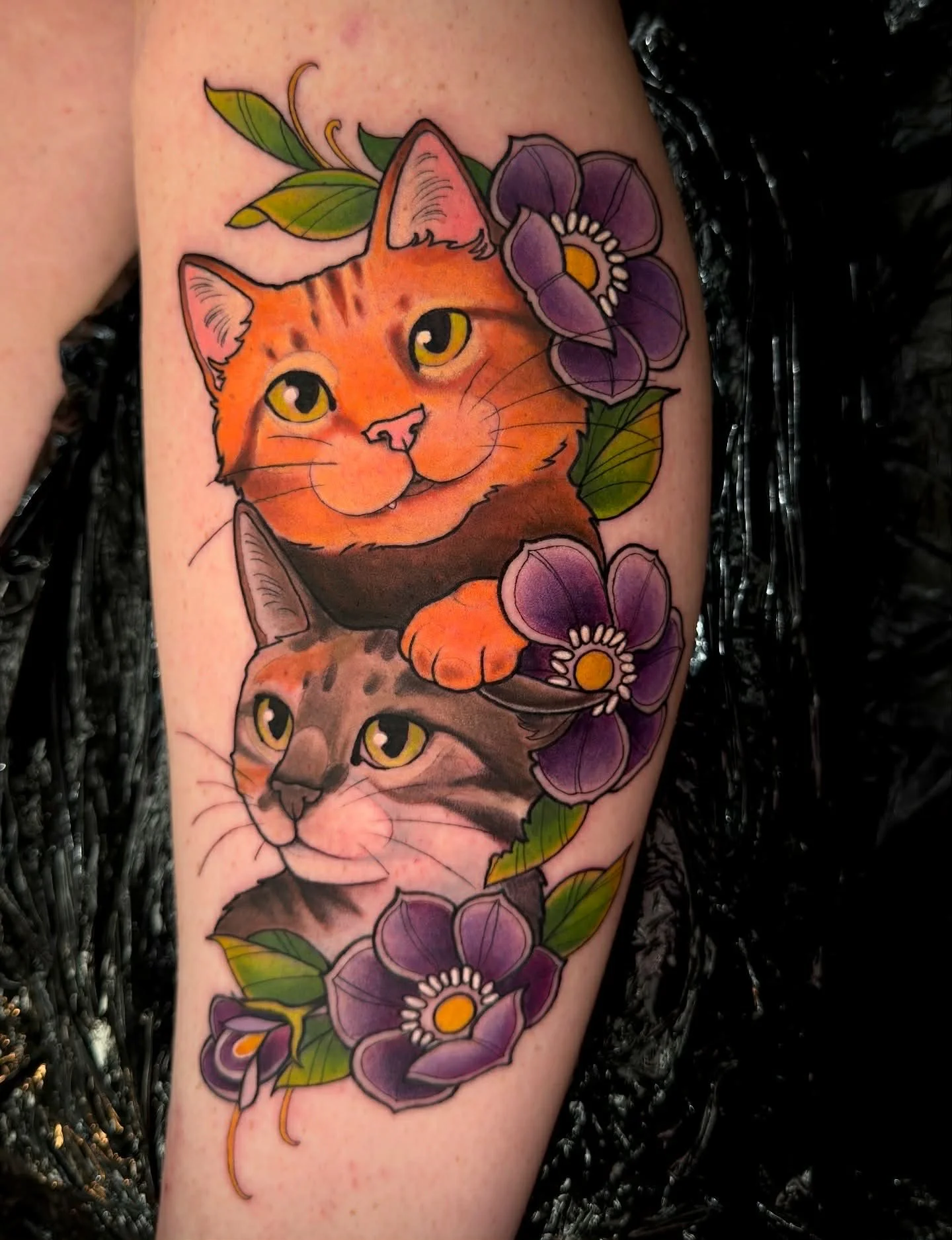 A colorful tattoo of two cats surrounded by purple flowers and green leaves on a person's arm.