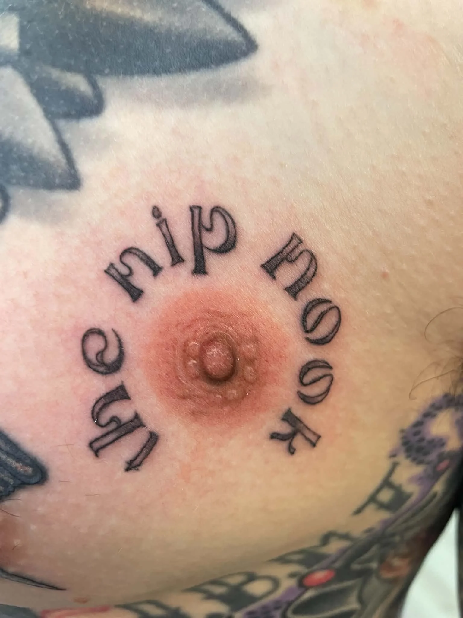 Close-up of a tattoo with black ink, featuring text in a curved arrangement on the skin, and a dark shaded design above.