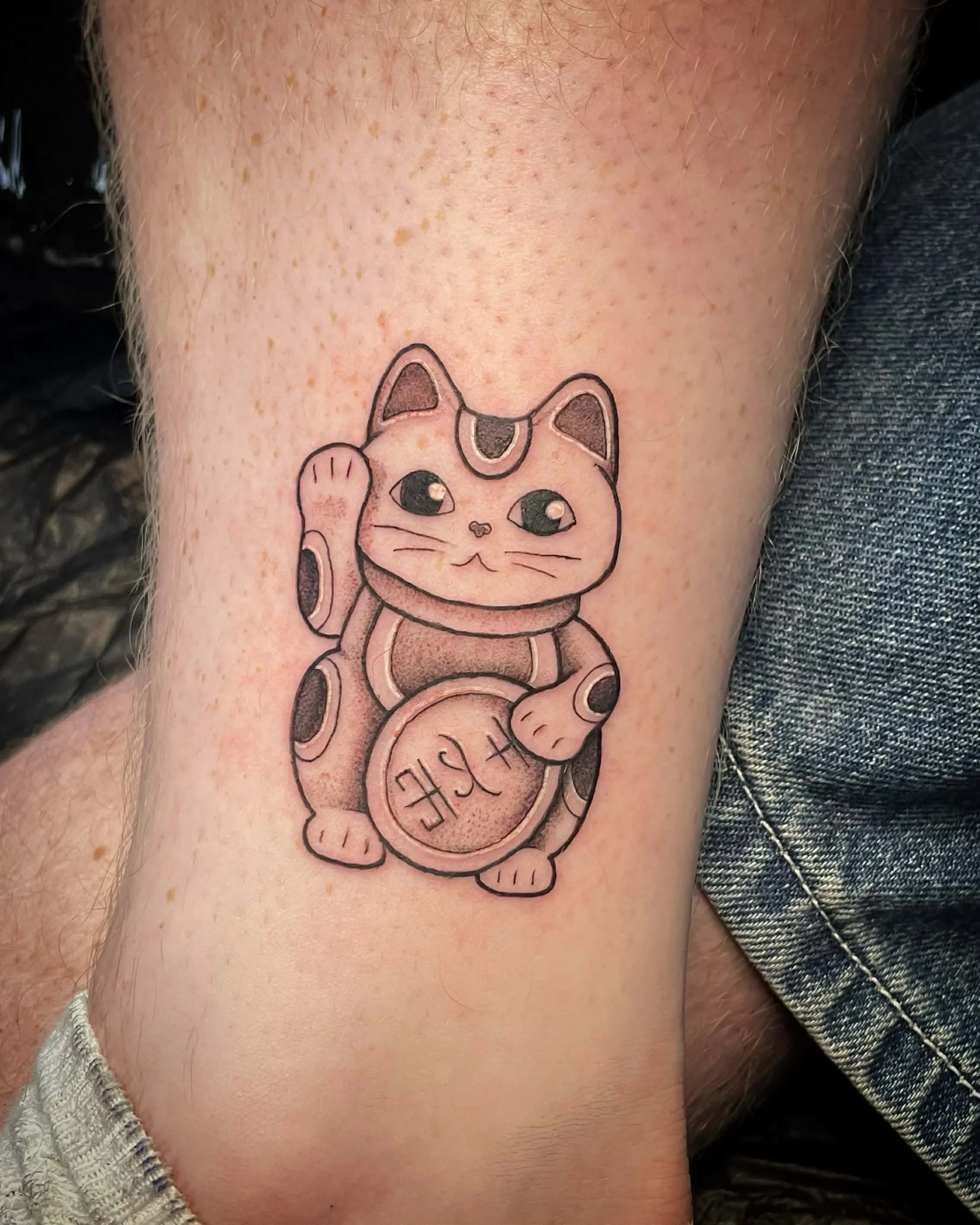Tattoo of a Maneki-neko, a Japanese lucky cat, with a raised paw, holding a coin with some characters, depicted in black and gray ink.