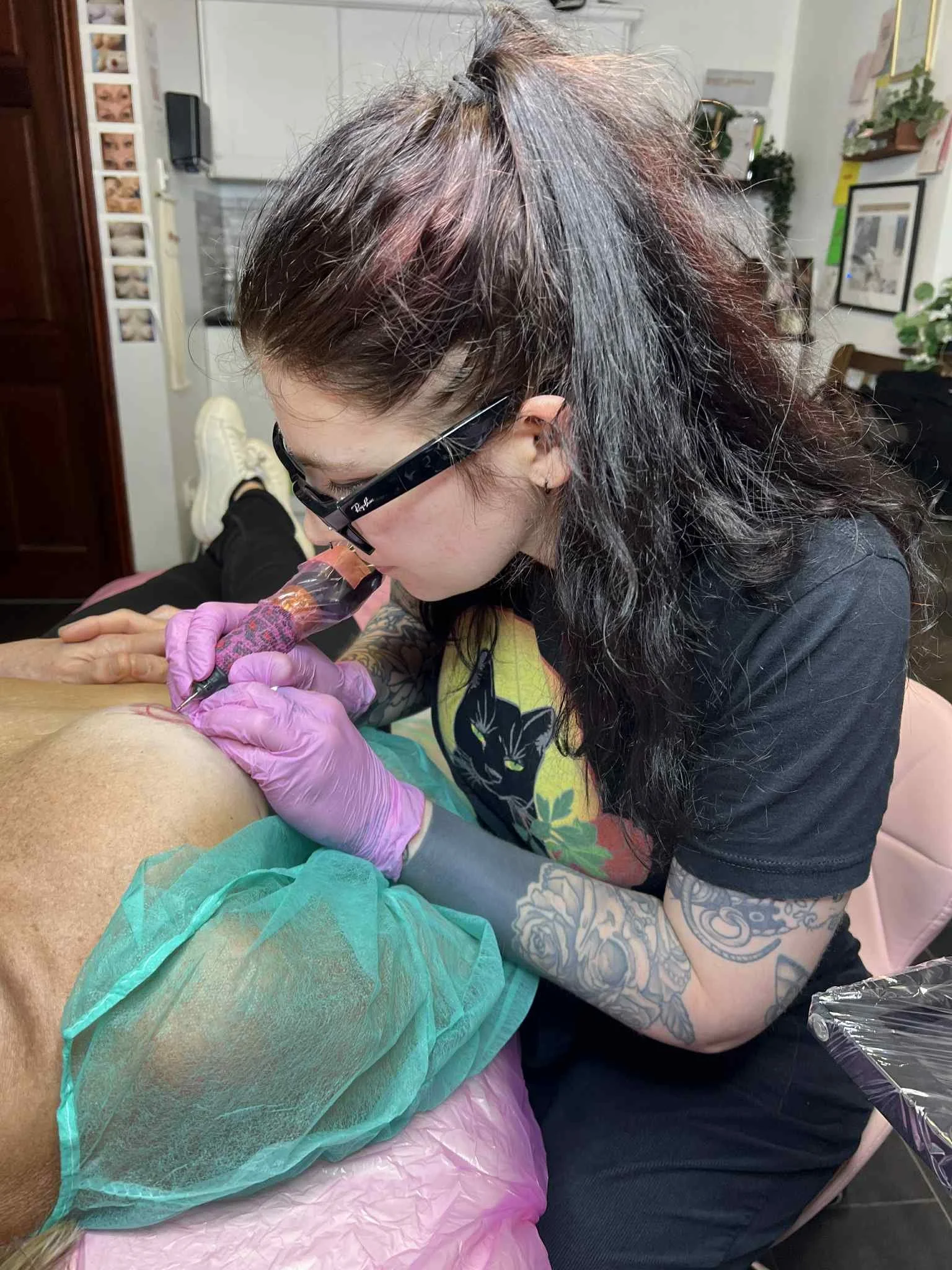 Tattooed woman with glasses and pink gloves tattooing a restorative 3d Areola tattoo in a tattoo studio.