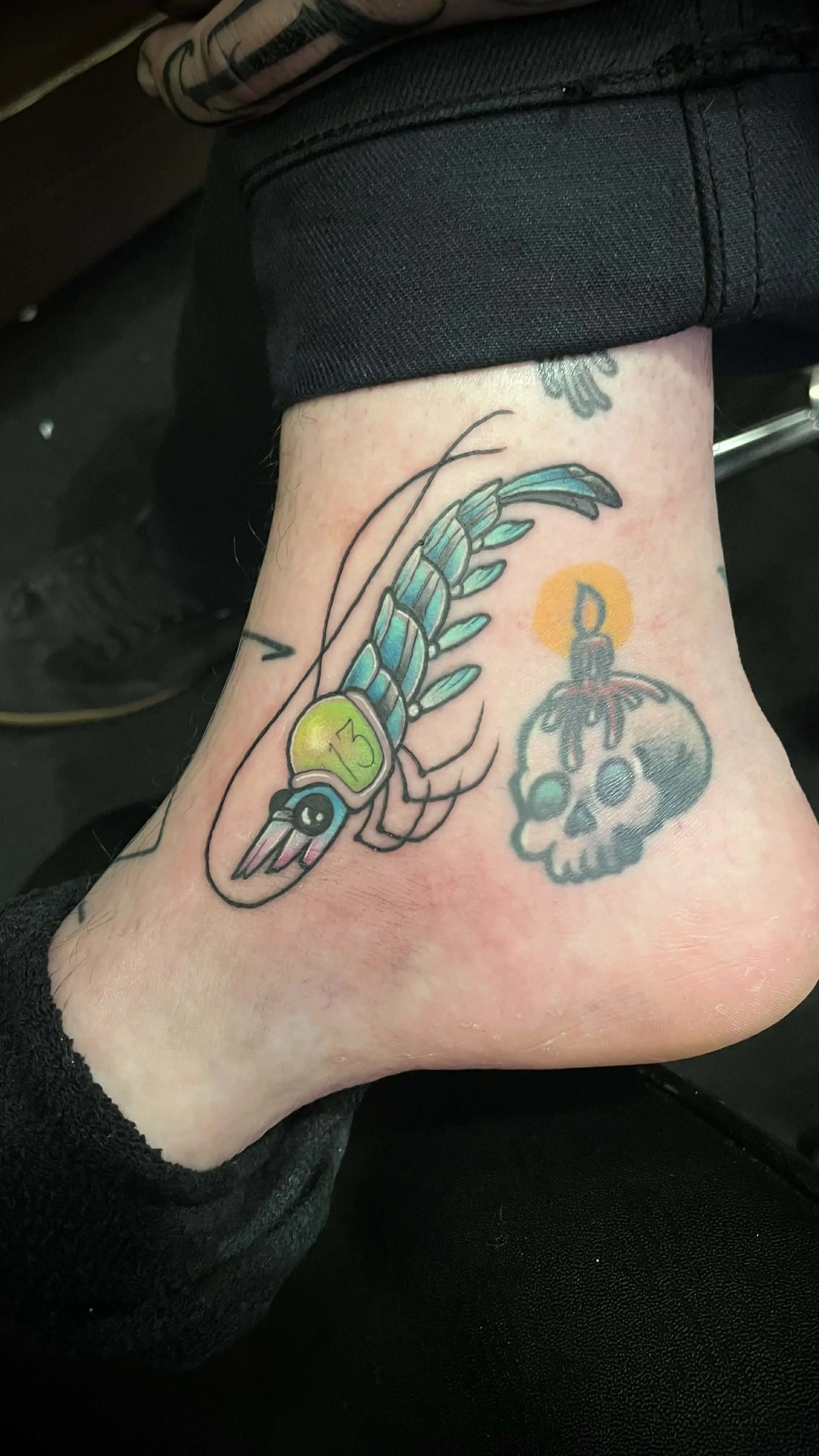 Tattoo of a shrimp with a digital watch displaying 13 on its shell, and a skull with a lit candle on top.
