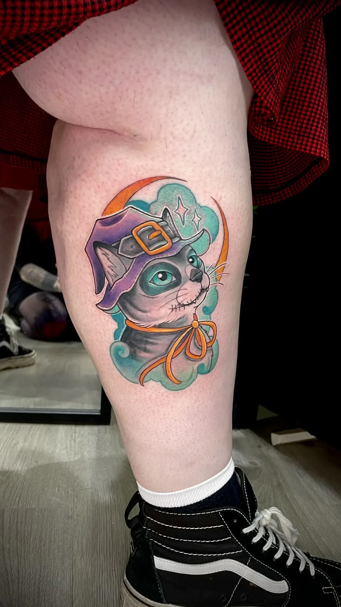 A colorful tattoo of a smiling cat dressed as a witch, wearing a purple hat with a black band and buckle, and an orange ribbon around its neck, surrounded by mystical clouds and stars.