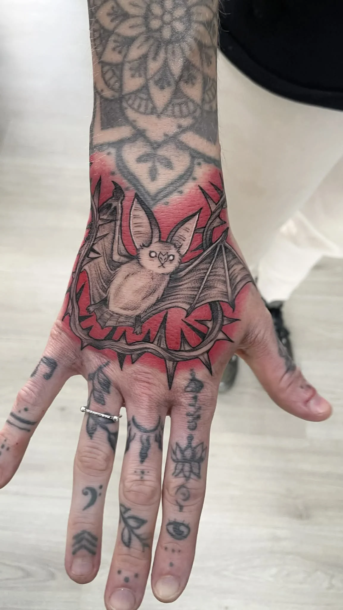 Tattoo of a bat with a cute face, wings spread, claws, and a background of red shading, covering the back of a hand and wrist.
