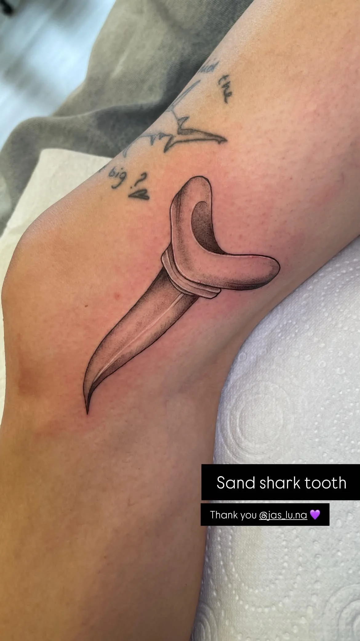 Tattoo of a sand shark tooth on a person's upper arm, with a bandage nearby and text indicating it is a sand shark tooth.