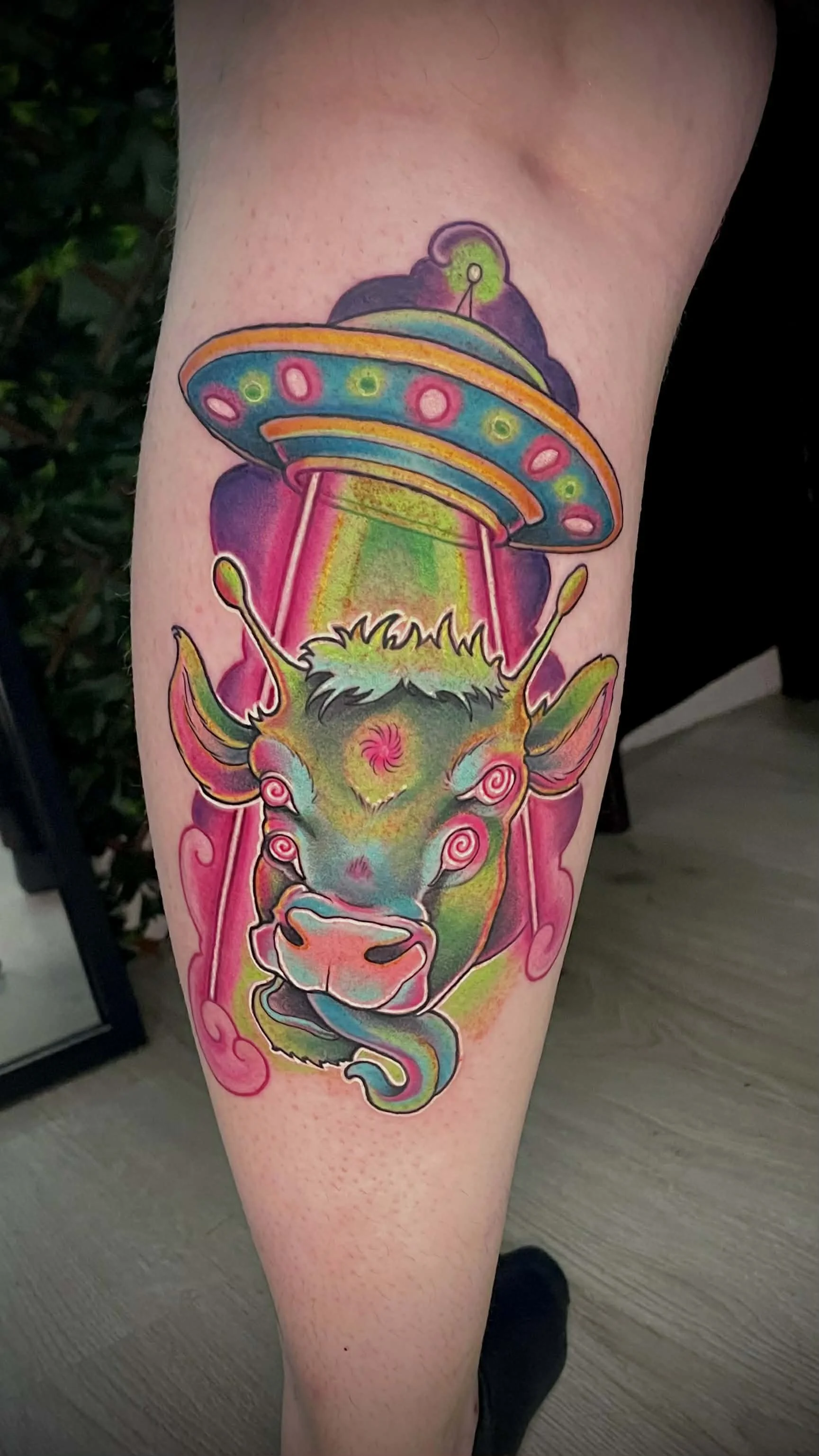 Colorful neon-style tattoo of a cow with hypnotic eyes, with a flying saucer and UFO above its head.