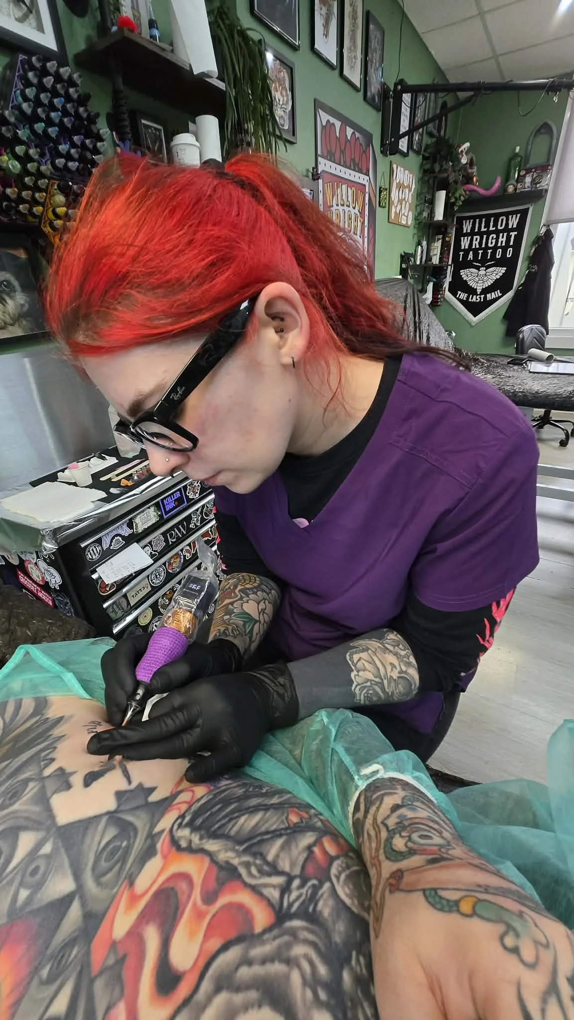A tattoo artist with red hair and glasses is tattooing a person's arm in a tattoo studio. The studio has green walls, framed artwork, and tattoo supplies visible in the background.