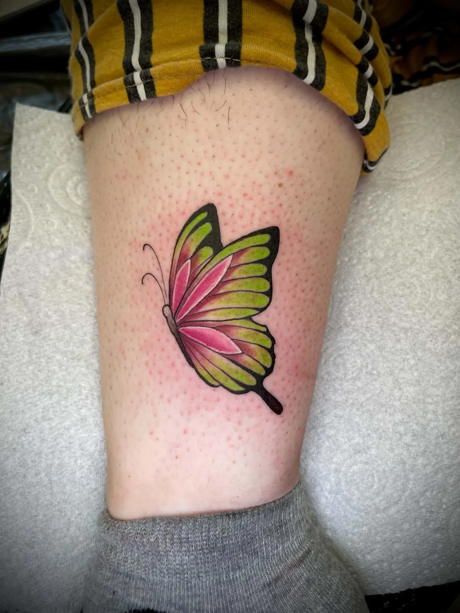 A colorful butterfly tattoo on a person's thigh, with emphasis on pink, green, black, and yellow colors.