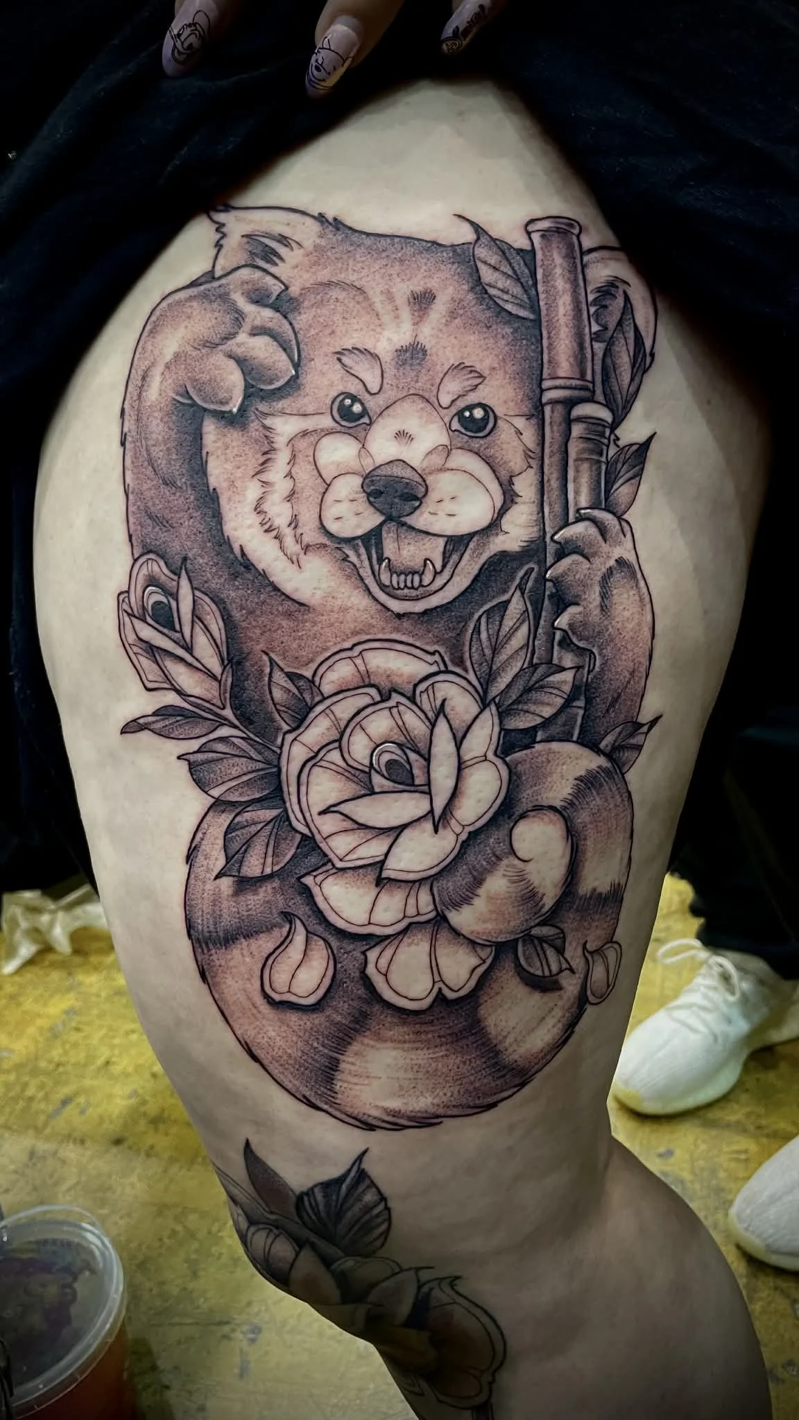 Tattoo of a red panda holding a bamboo stick, with leaves and flowers around it, depicted in a black and grey style on a person's thigh.