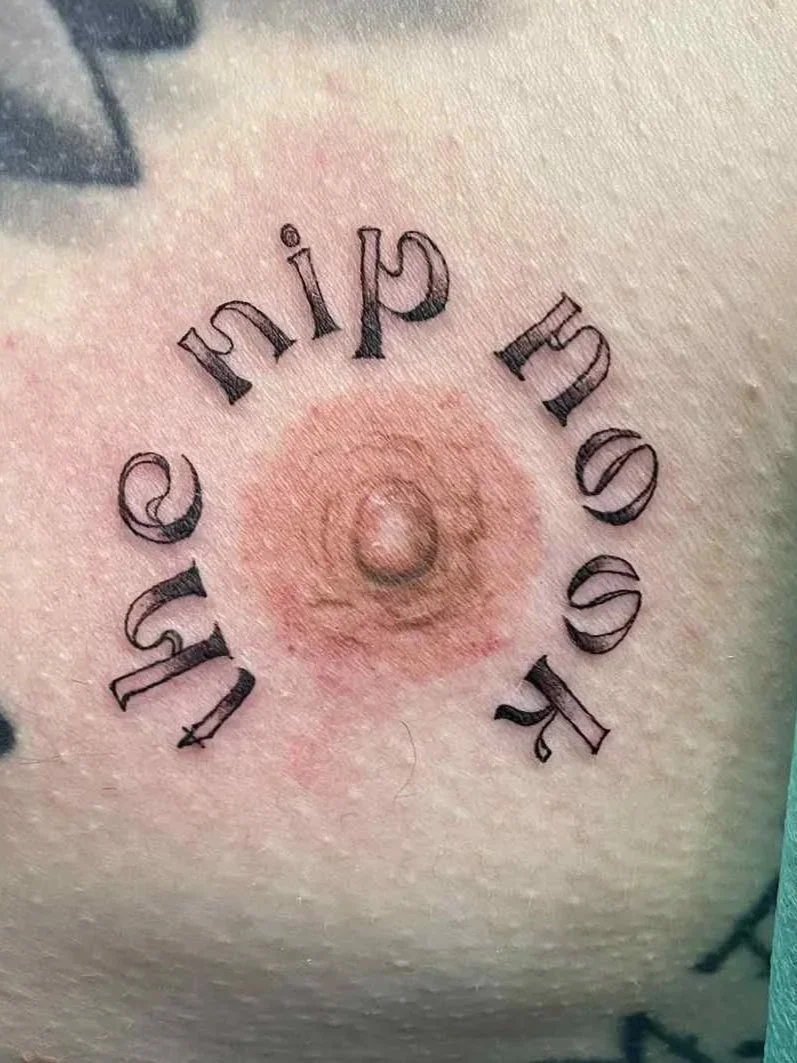 Tattoo of the words 'the nip nook’ arranged in a circle around a 3d realistic areola nipple tattoo on flat skin.