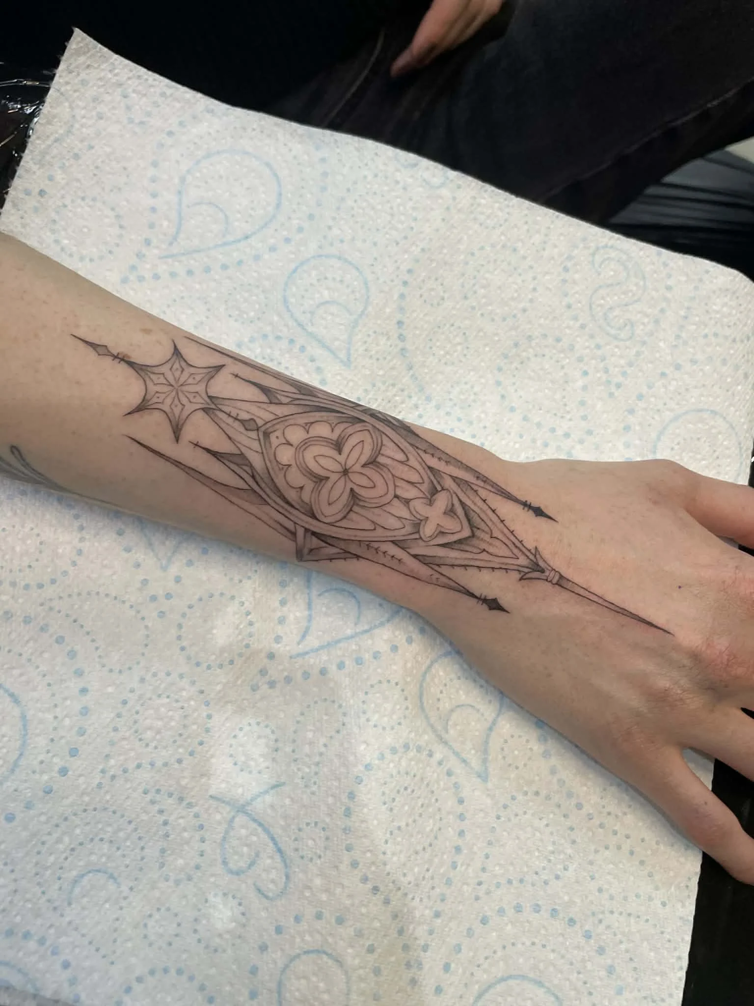 A black ink tattoo of a decorative cathedral, gothic archway design on a person's forearm, with a disposable tattoo cover sheet underneath.