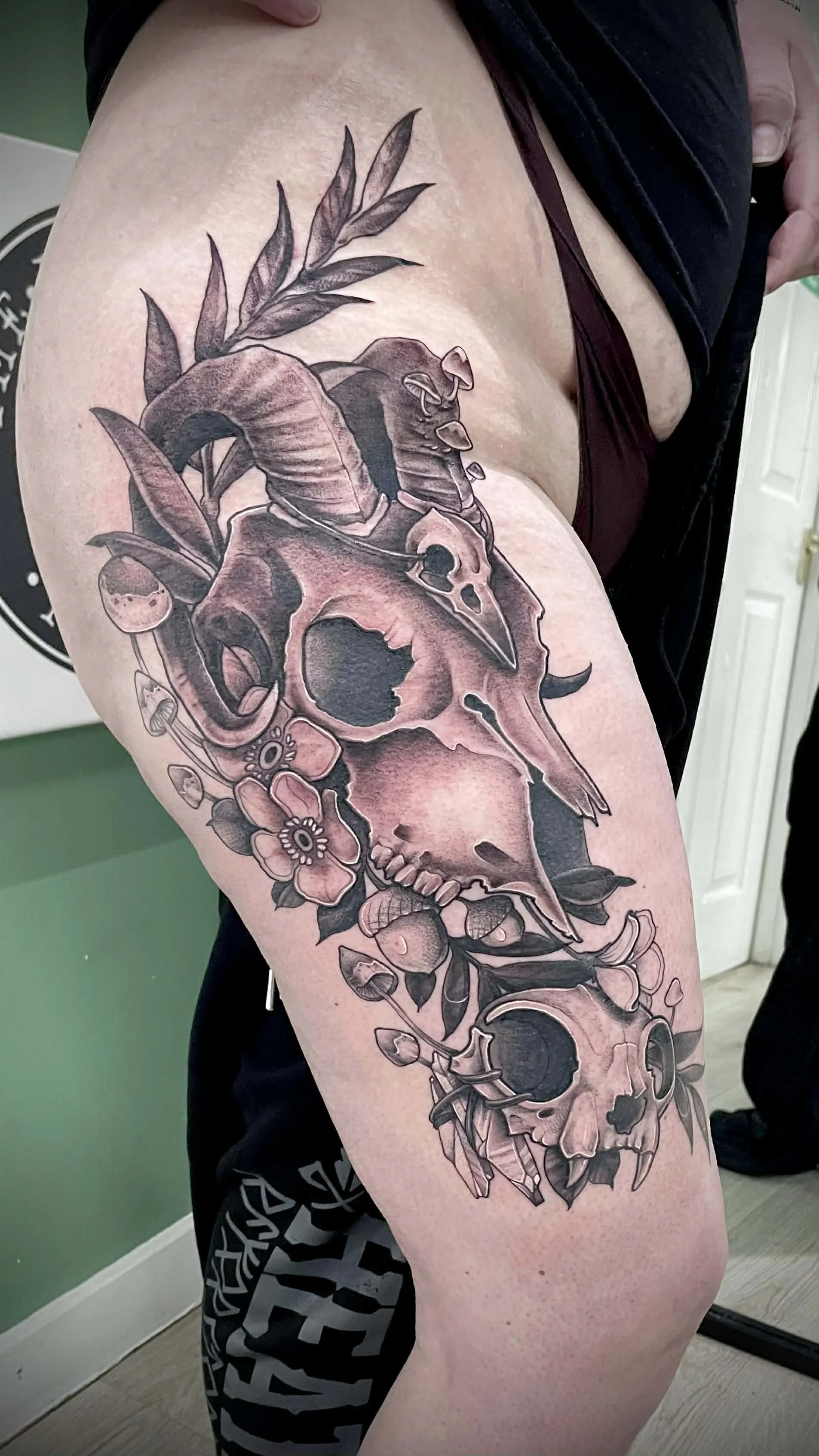 A realistic black and gray tattoo of two skulls with leaves, flowers, and other botanical elements on a person's upper arm.