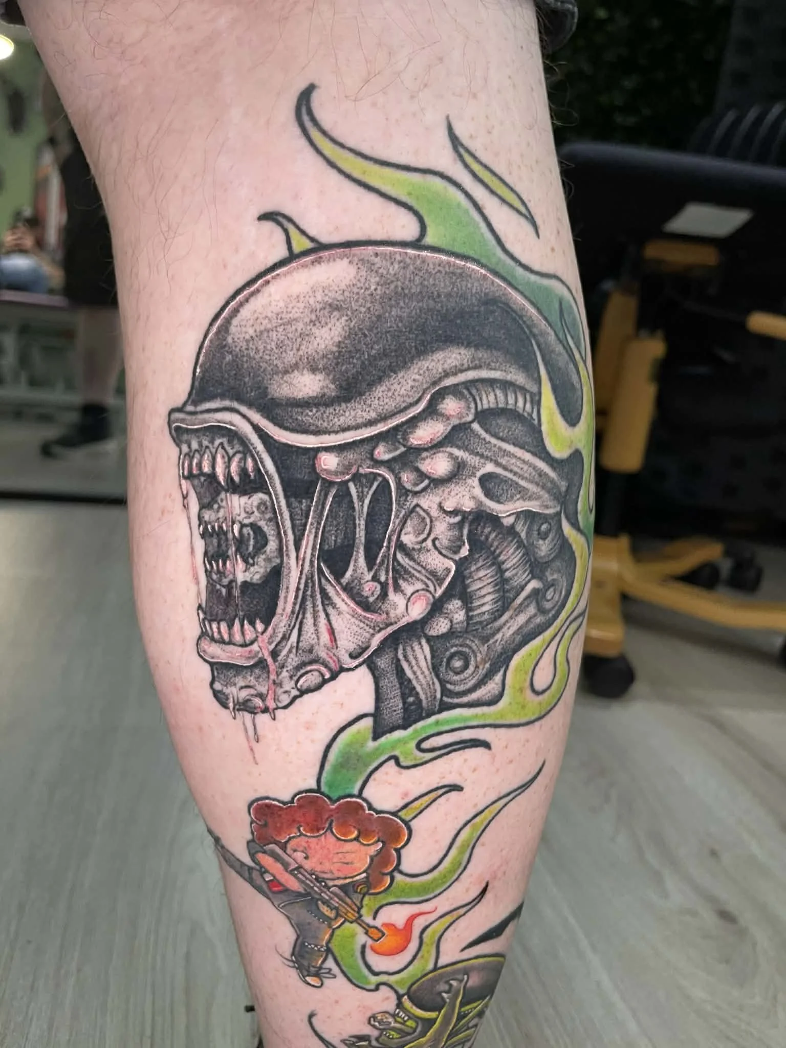 Tattoo of a screaming alien creature with an elongated head and exposed brain, surrounded by green and yellow flames, with a small cartoon figure playing an electric guitar emitting flames. Alien tattoo, Ellen ripley, xenomorph jonesy